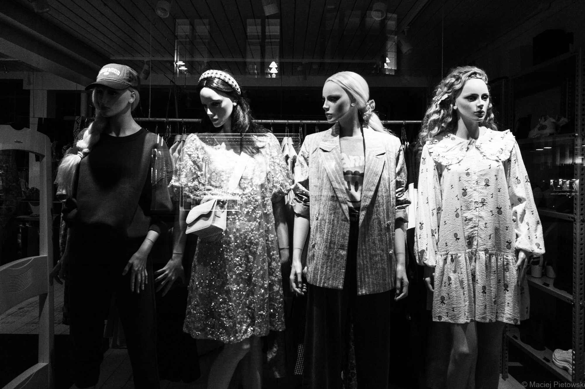 Four mannequins dressed in fashionable clothing on display in a retail store window, with racks of clothes visible behind them and the store interior reflected on the glass.