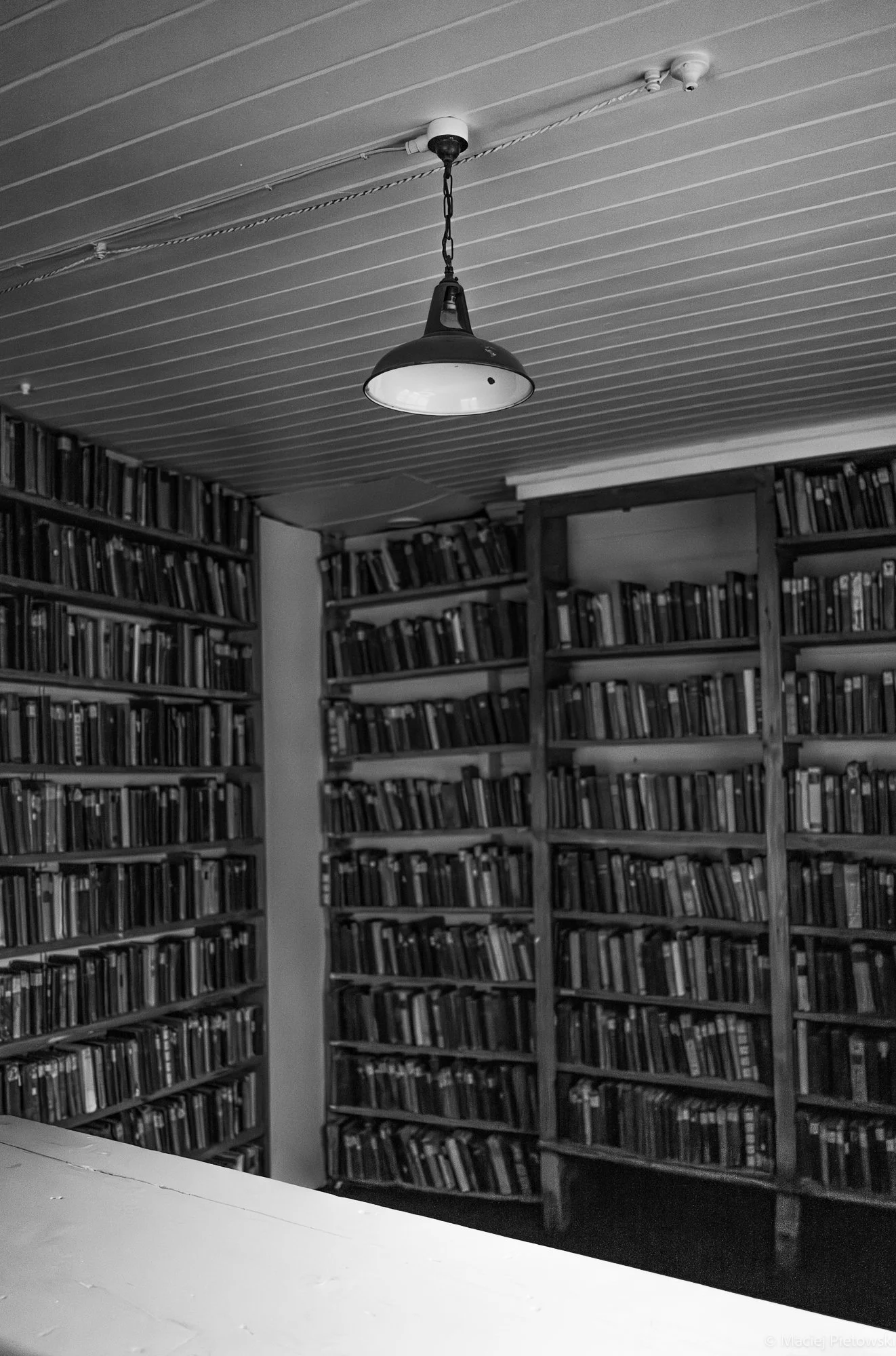 Black and white photo of a library with bookshelves filled with books, a ceiling with horizontal wooden panels, and a hanging industrial-style light fixture in the center.