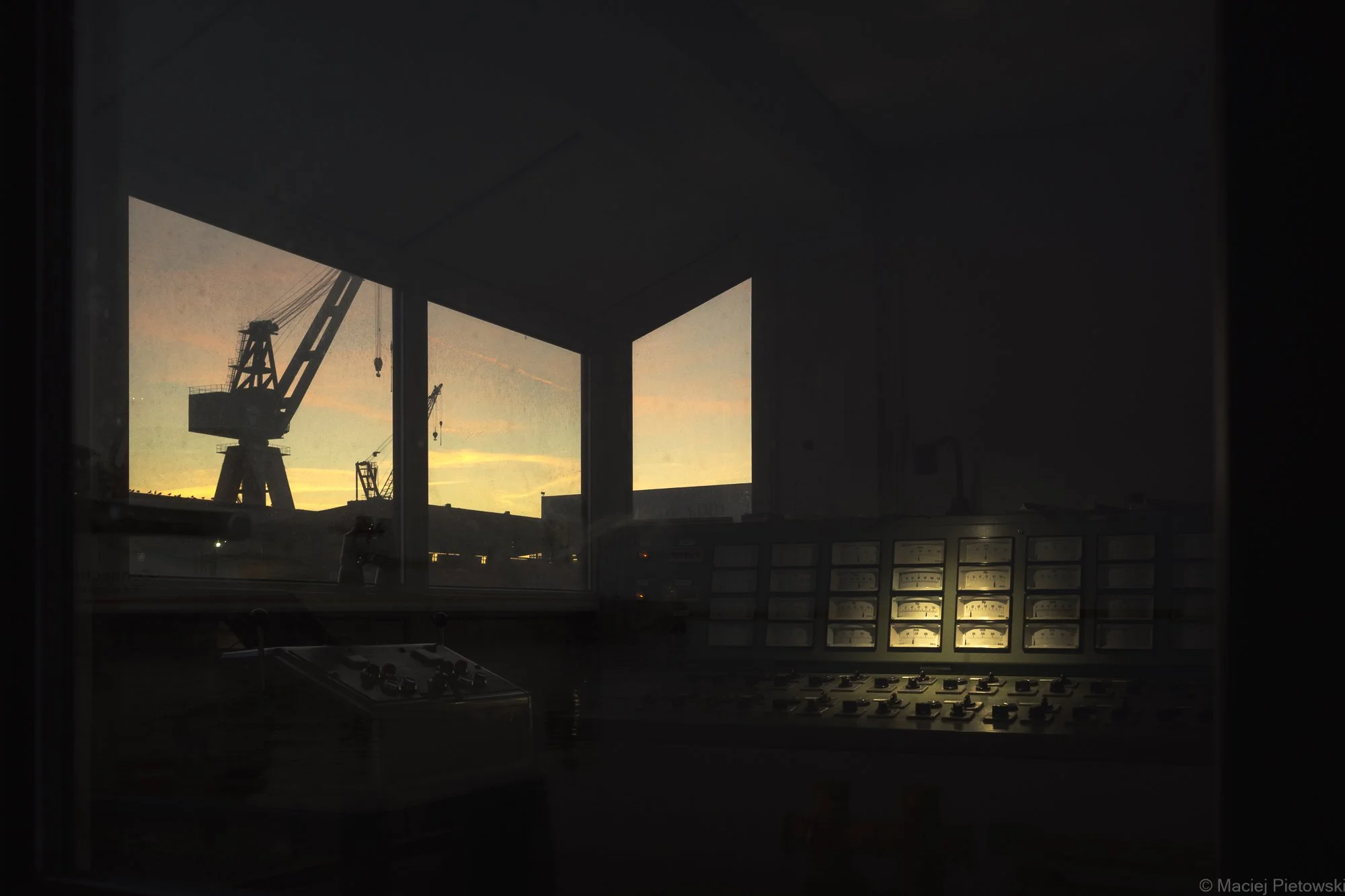 Interior of an control room with a large window showing a construction crane and sunset sky. Inside the room, there are control panels with numerous buttons and gauges, and a desk with equipment.