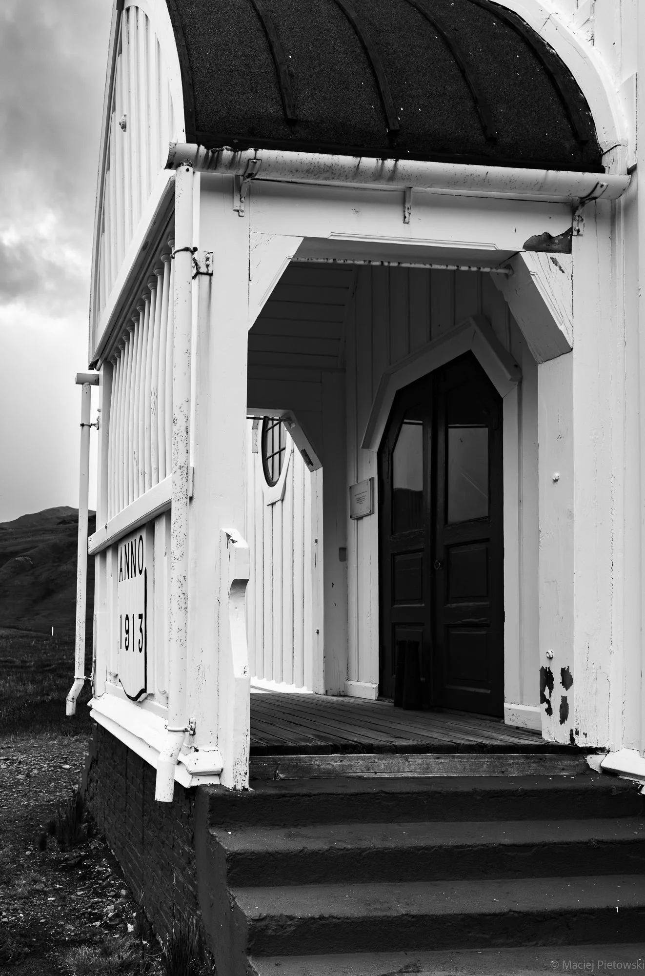 Black and white photo of the entrance to a small church or chapel with a curved roof and a black door, built on a raised platform with steps, set in a rural landscape with rolling hills and cloudy sky in the background.