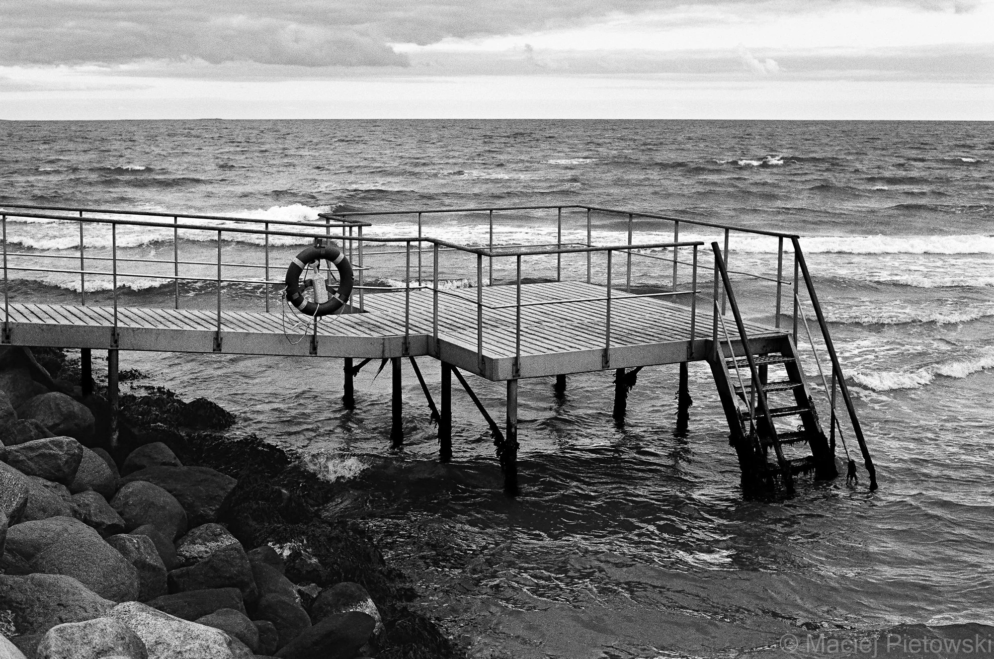 A black and white photo of a seaside wooden pier with metal railings and a life preserver hanging on a post. The pier extends over rocks into the ocean with waves, and a cloudy sky is above.