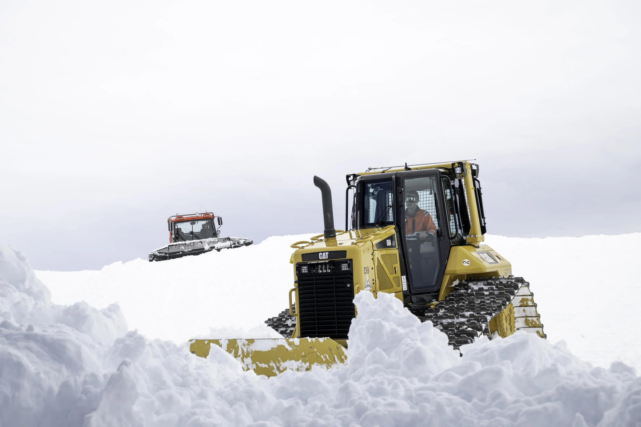 Two workers in orange safety jackets and helmets operate yellow snow removal machines with tracks on a snowy landscape. The sky is overcast.