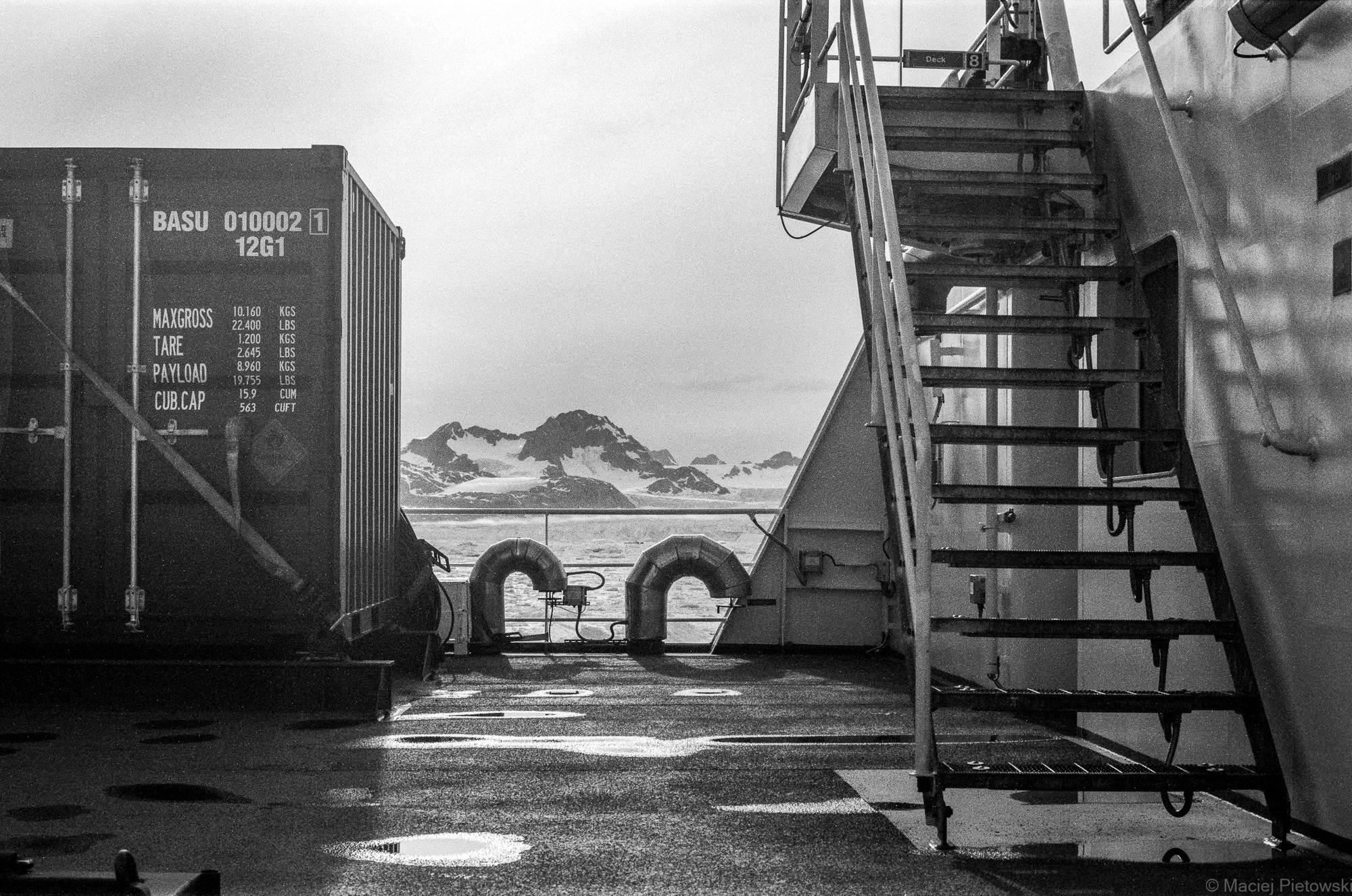 View from a ship deck showing stairs on the right, a container on the left with maritime information, and snowy mountains in the distance under a cloudy sky.