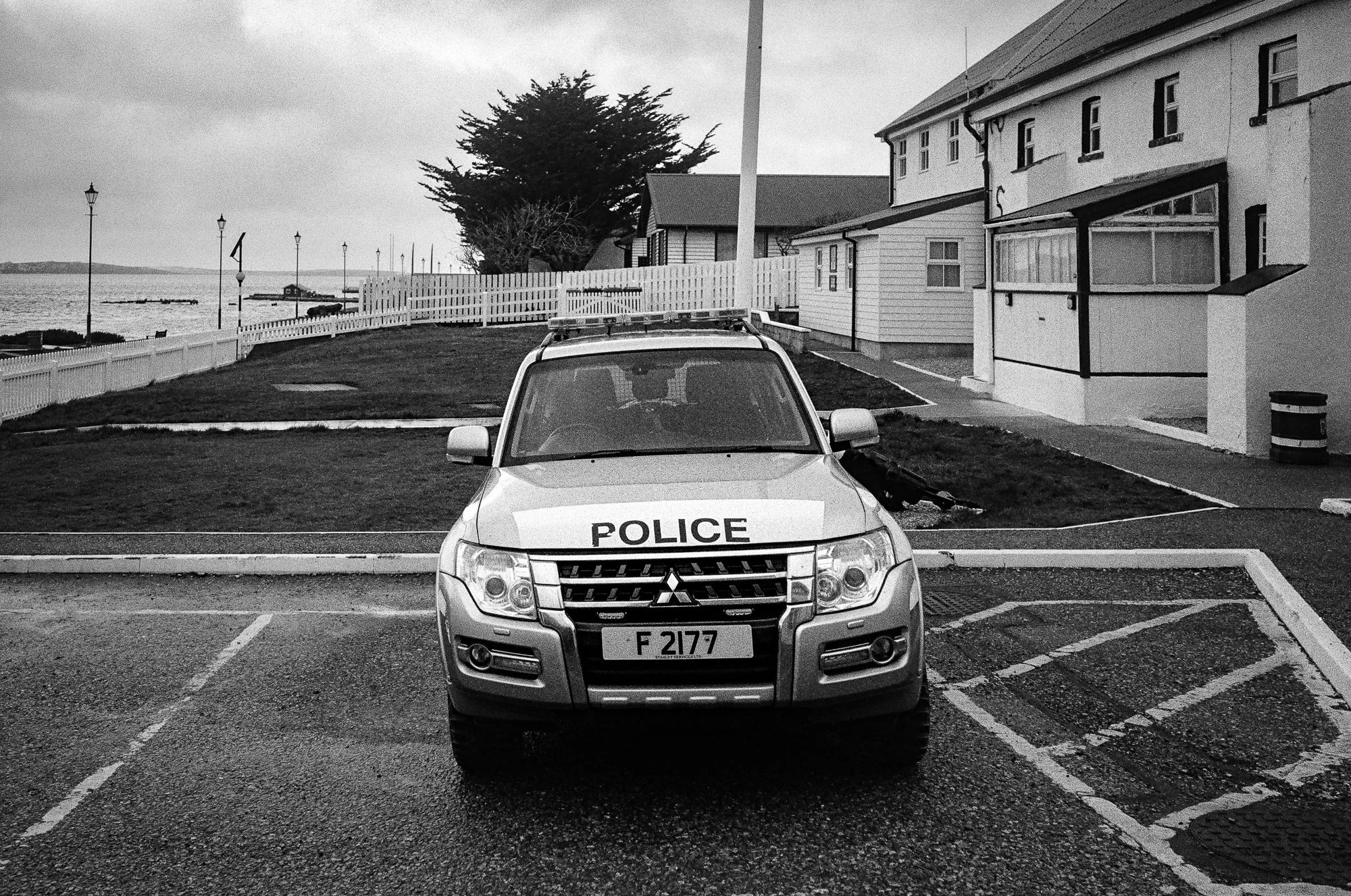 A police vehicle parked in a lot facing forward, with a license plate that reads 'F 2177'. In the background, there are beachside buildings, a white picket fence, a large tree, and a view of the ocean with a cloudy sky.