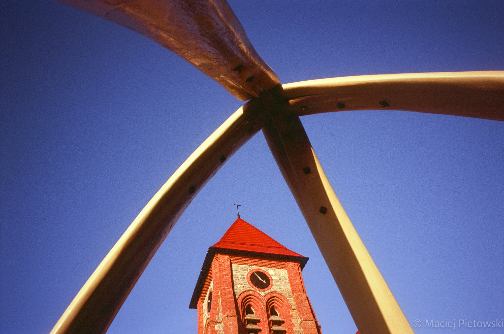 A low-angle view of a red brick church tower with a clock, framed by curved wooden beams or sculptures against a clear blue sky.