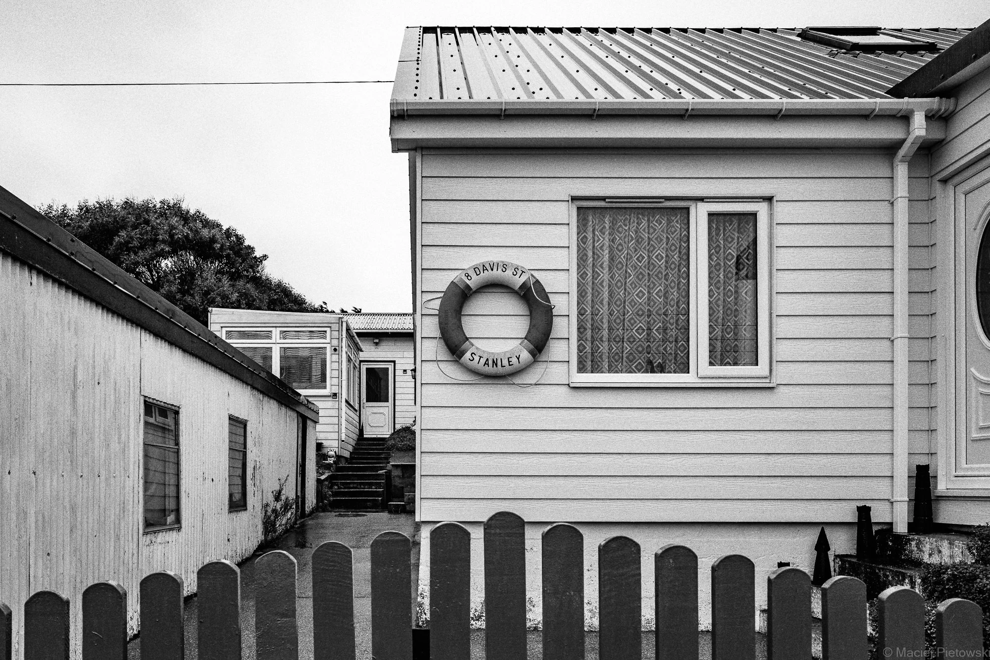 Black and white photo of a house with a corrugated metal roof and siding. A round life preserver with the text '8 DAVIS ST STANLEY' hangs on the exterior wall. A fence in the foreground, a set of stairs leading to a door at the back of the house, and