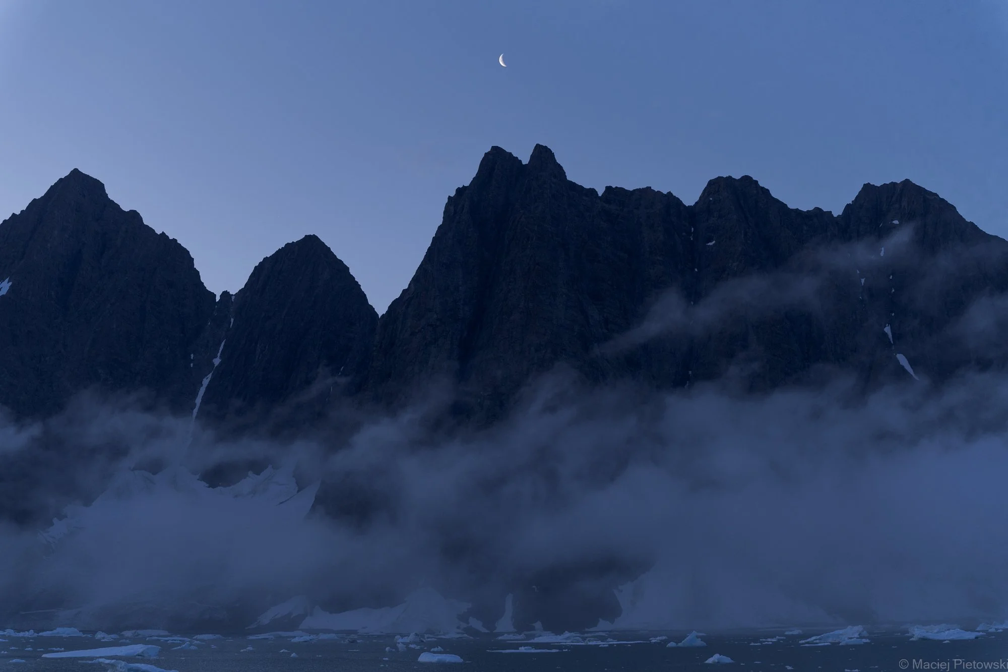 Mountain peaks with snow, a crescent moon in the sky, and fog or low clouds at the base of the mountains.