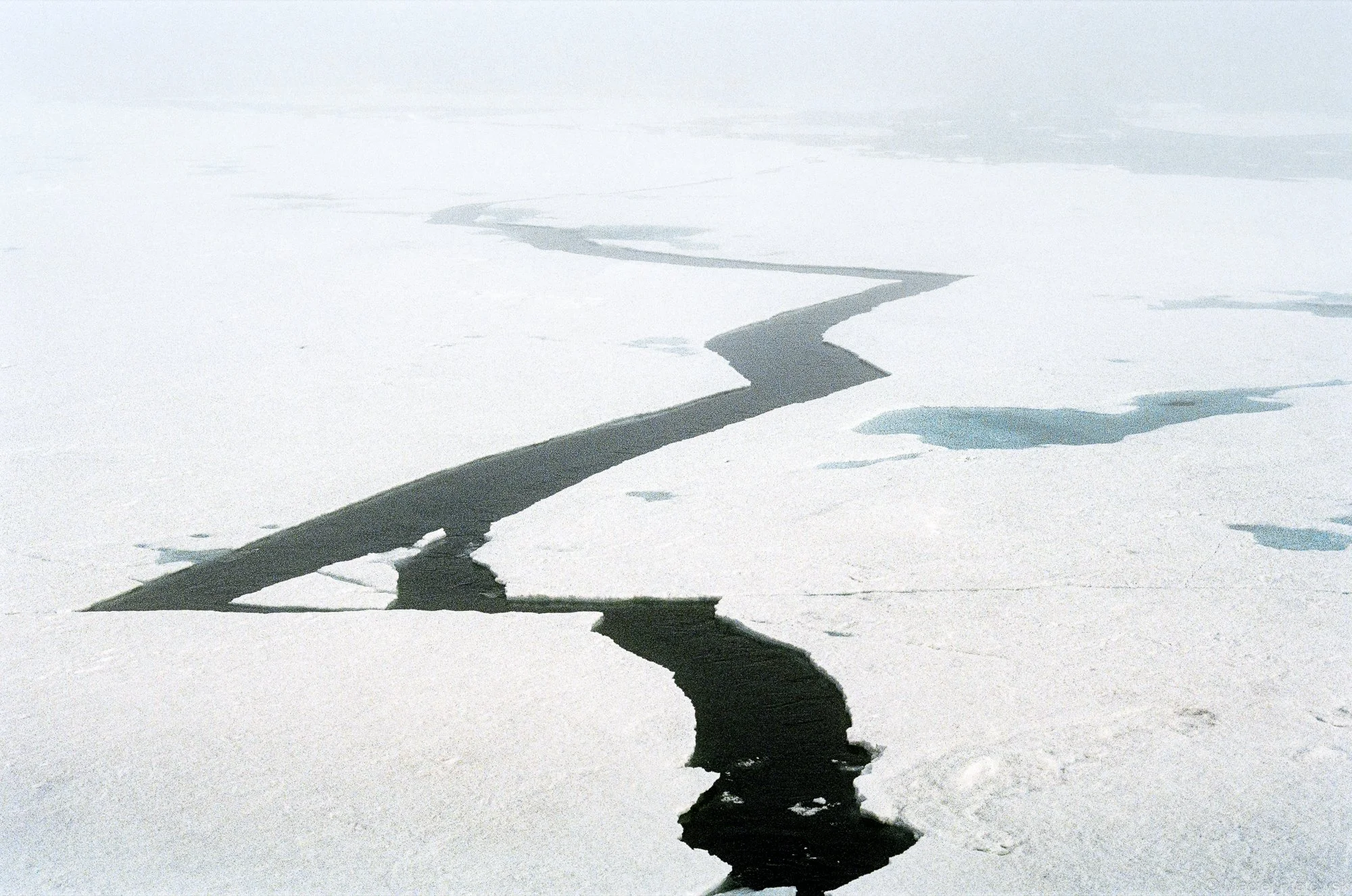 A vast snow-covered landscape with a dark, winding crack or river cutting through the ice, extending into the horizon under a foggy or overcast sky.