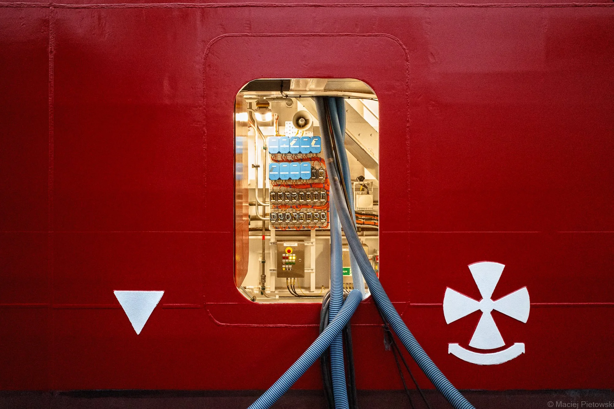 View of electrical components and cables through a rectangular opening in a red panel with white arrow and sun symbols.
