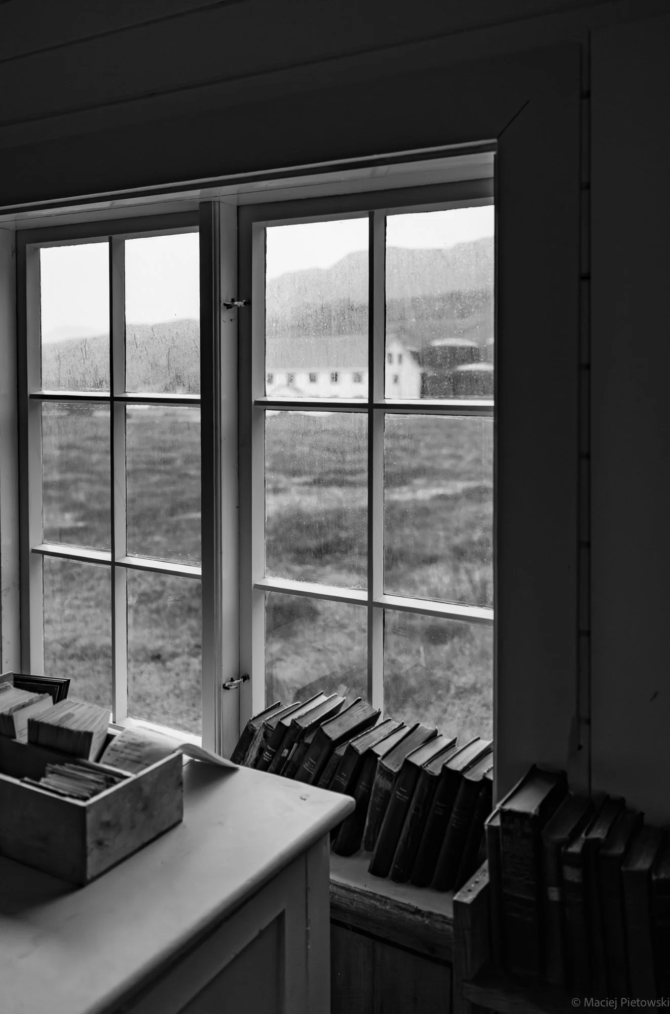 Black and white photo of a window with a view of mountains and buildings outside. Inside, a desk with a container of books and a row of books on the windowsill.
