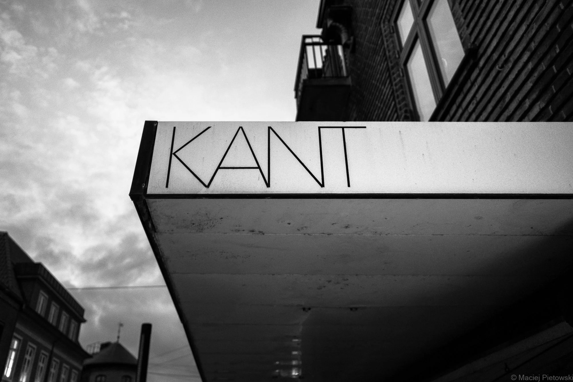 Black and white photograph of a building's sign that reads 'KANT' with a building with windows and a balcony on the upper floors in the background.