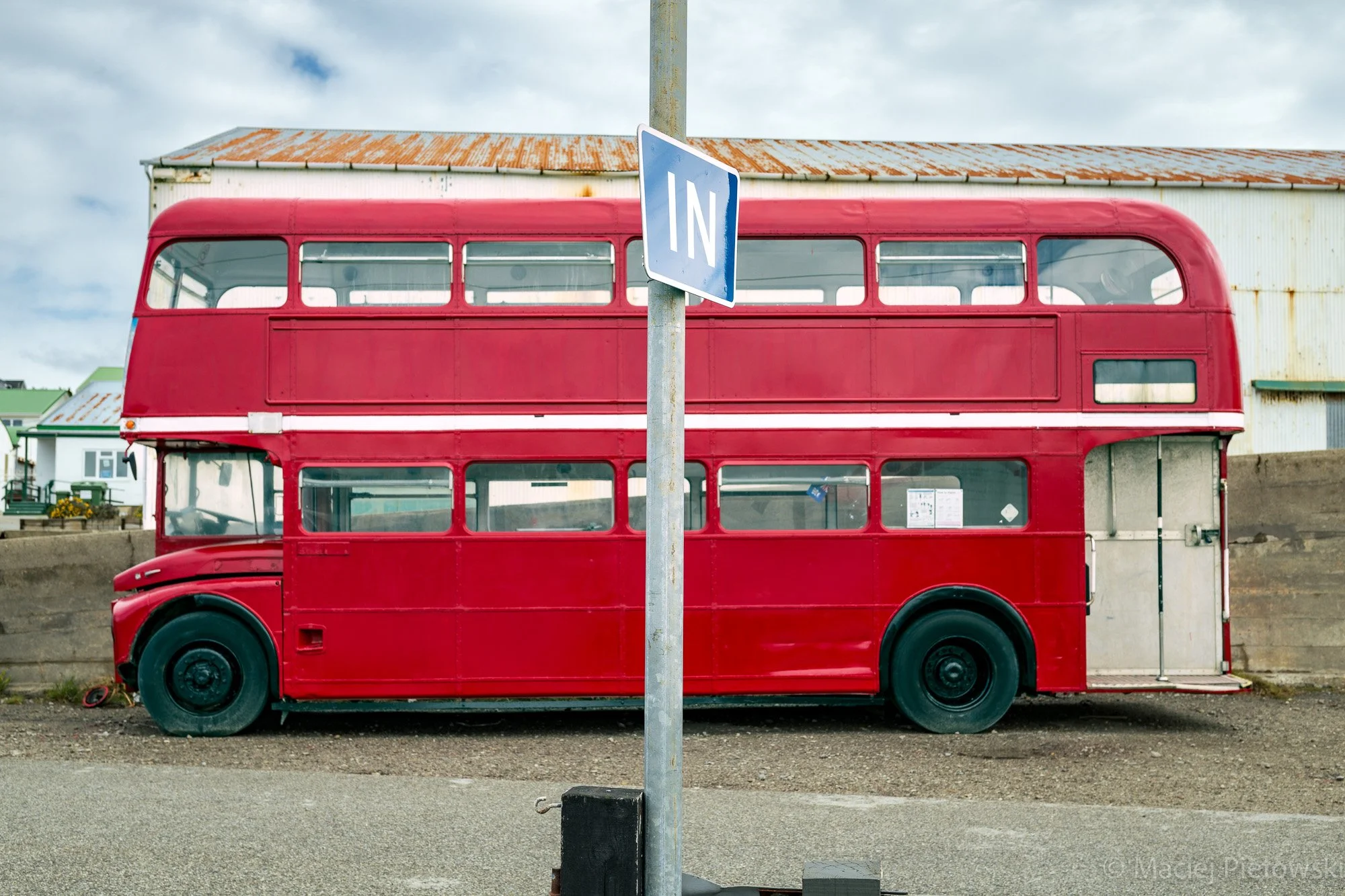 A red double-decker bus parked behind a blue 'IN' traffic sign on a city street with cloudy skies overhead.