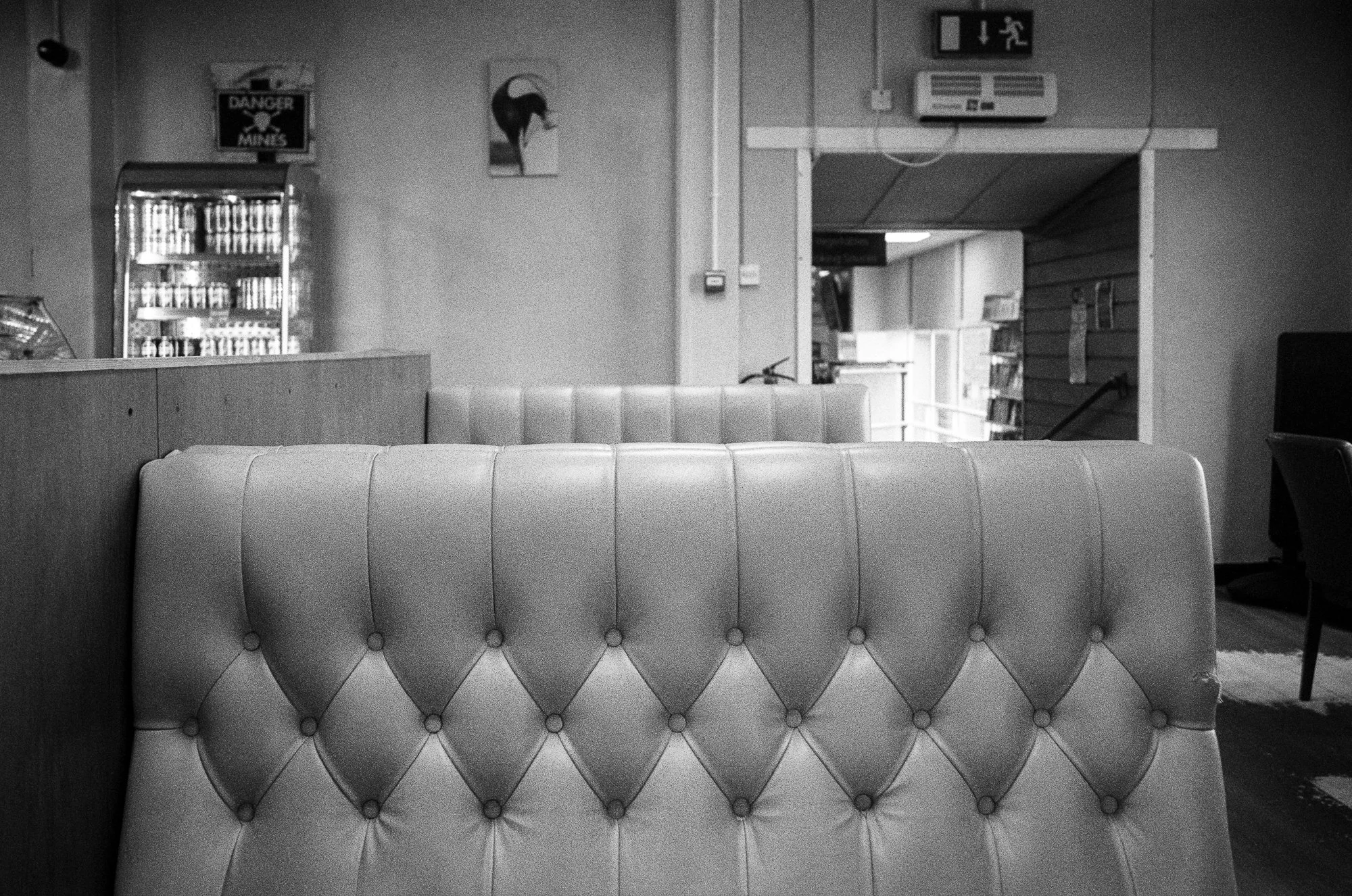 Black and white photo of an indoor space with upholstered furniture in the foreground, a vending machine to the left, artwork on the wall, and an opening leading to another room with shelves on the right.