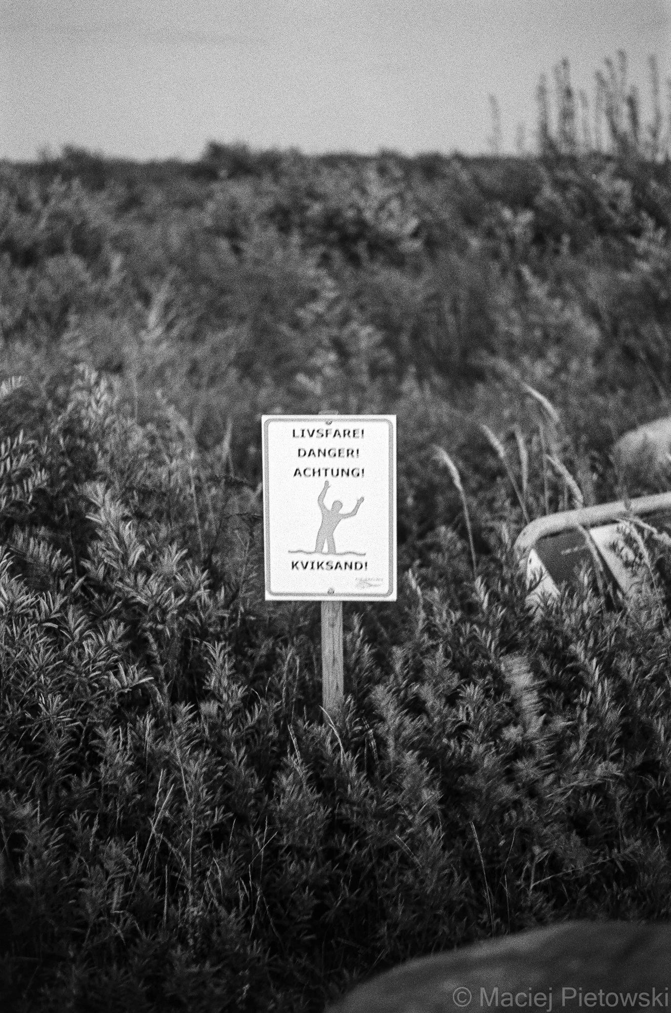 A warning sign in front of a grassy field with a hill and sparse bushes or trees in the background. The sign depicts a person falling backwards with arms raised, indicating danger. The text on the sign reads, "LIVSFARRE! DANGER! ACHTUNG!" and "KVIKSA