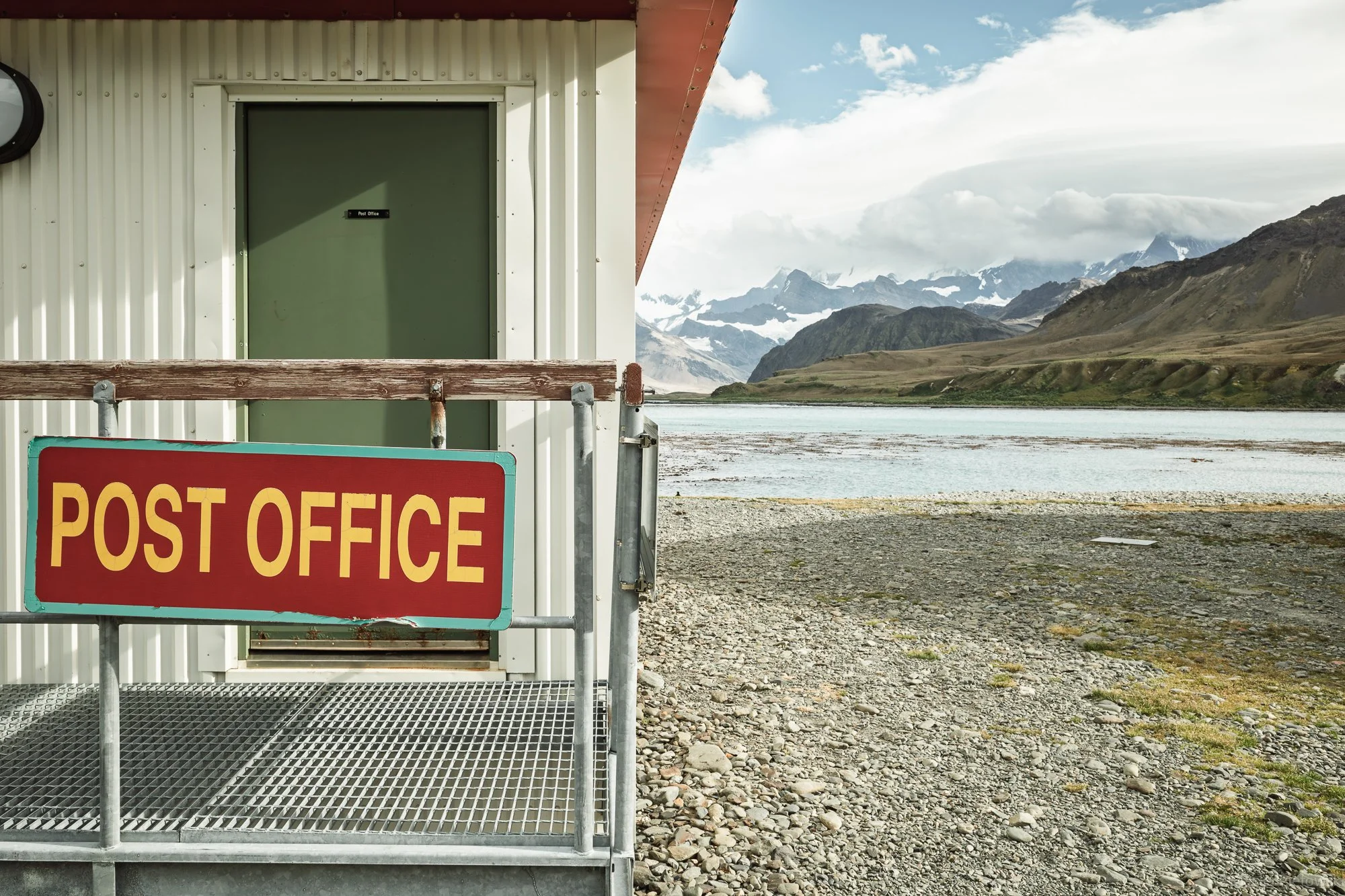 A small green post office building with white corrugated siding, next to a body of water and mountainous landscape under a partly cloudy sky.