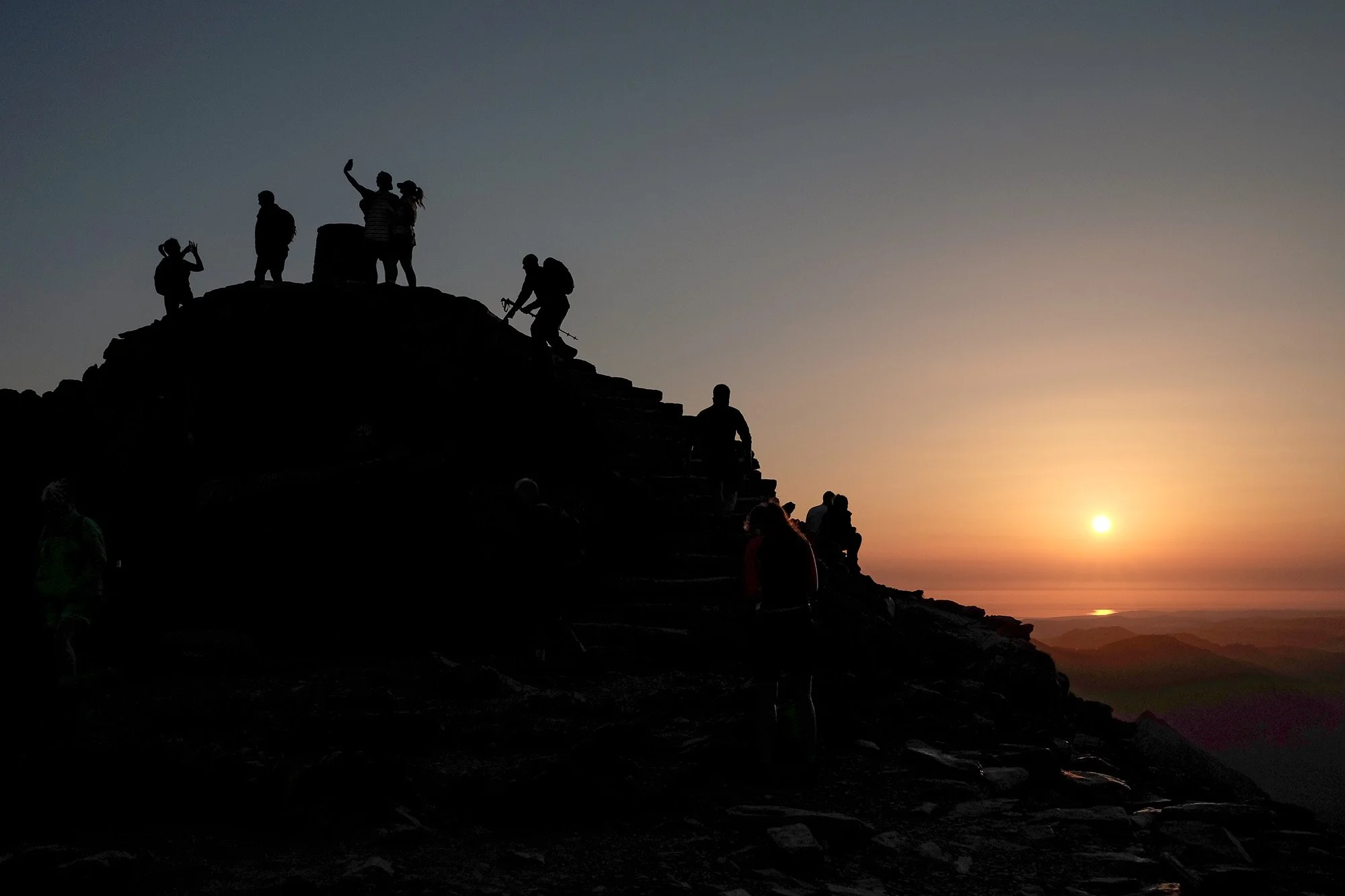 Silhouettes of hikers on a rocky ridge at sunset, some taking photos or resting, with a clear sky and the setting sun in the background.