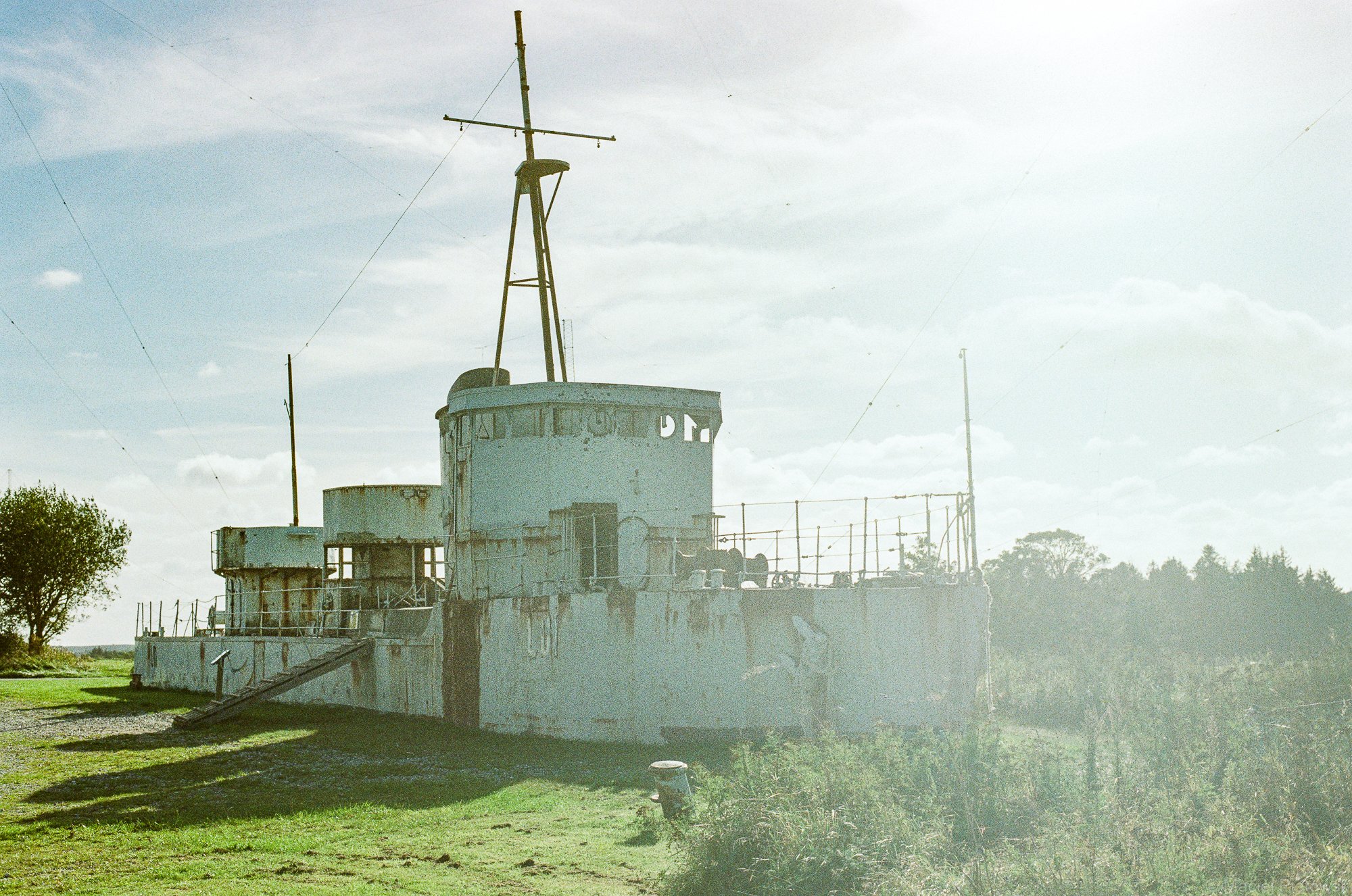 An old, abandoned military ship resting on land with some trees and grass around it, under a cloudy sky.