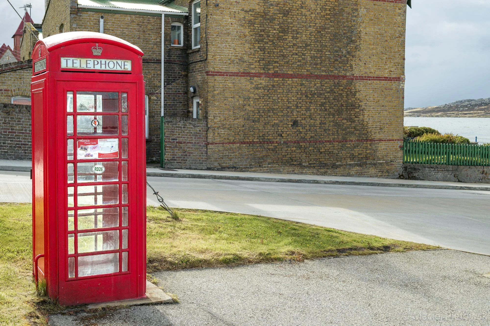 A traditional red British telephone booth on a sidewalk beside a brick building and a street, with grassy area in the foreground and a body of water in the background.
