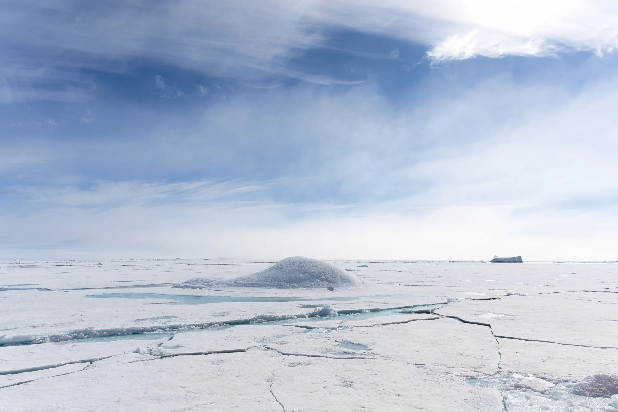 A vast icy landscape with cracks and a small snow mound, under a partly cloudy sky with wispy clouds and a distant iceberg.