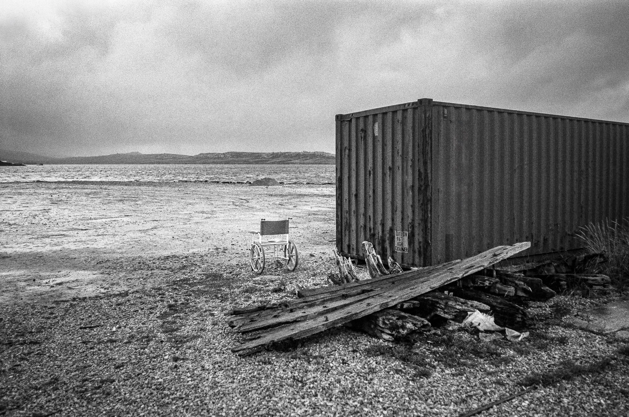 A black and white photograph of a beach scene with overcast skies, featuring a large shipping container, a wheelchair, a small fence, and some wooden debris.