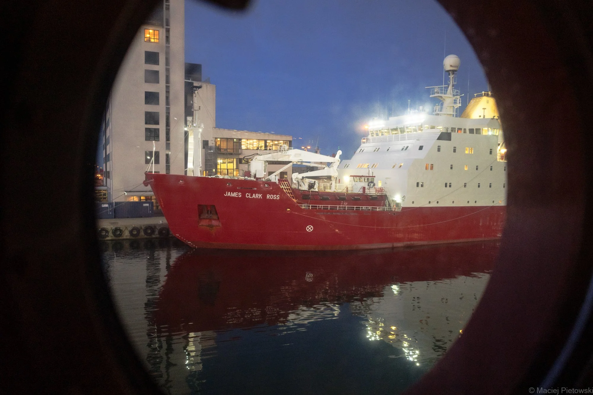 Photo of a large red and white ship named James Clark Ross docked at a modern harbor with buildings in the background, viewed through a round porthole.