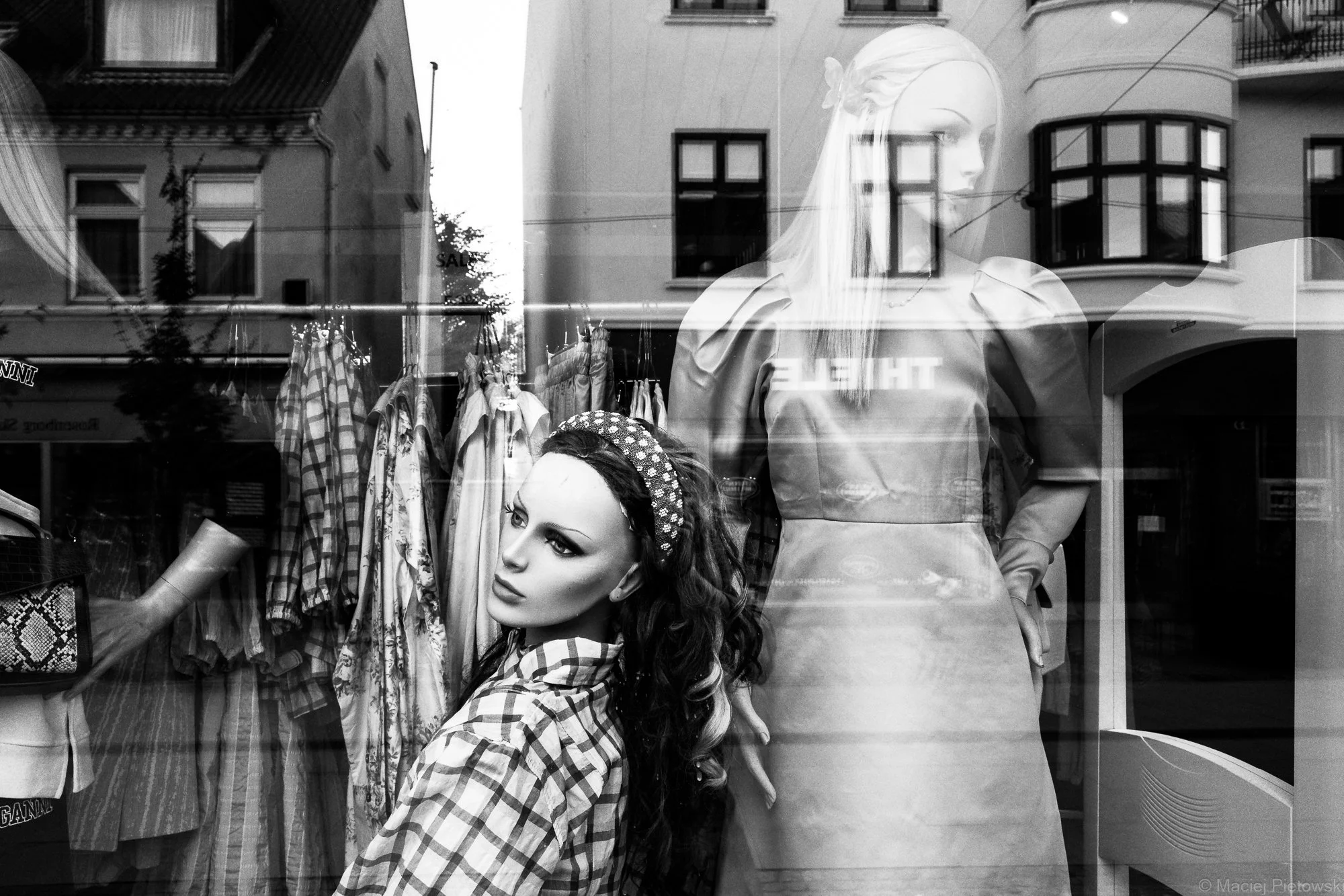 A store window display showing two mannequins dressed in stylish clothing. The reflection of buildings and the street outside is visible on the glass.