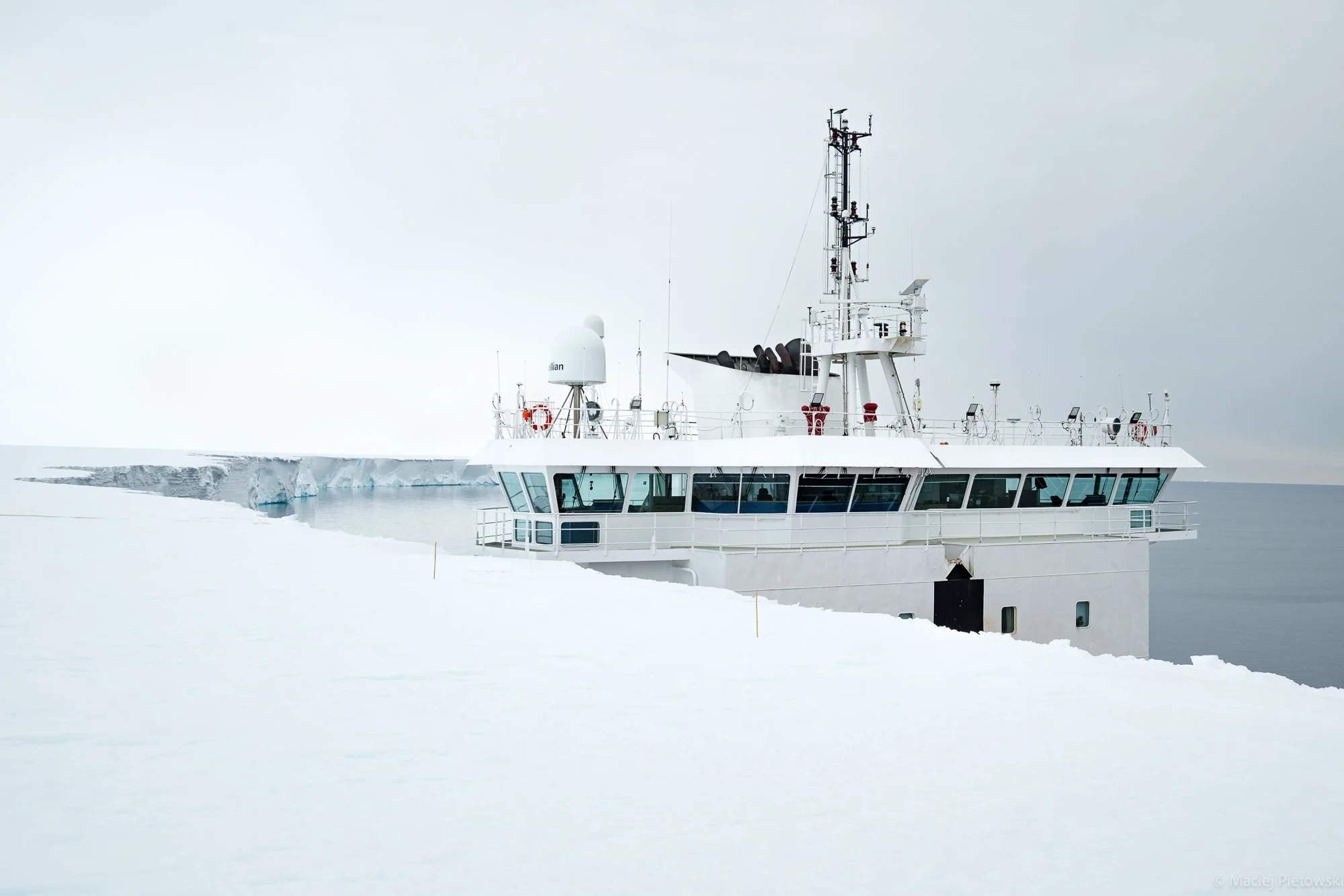 A white ship navigating through icy waters with snow-covered landscape and icebergs surrounding it, under an overcast sky.