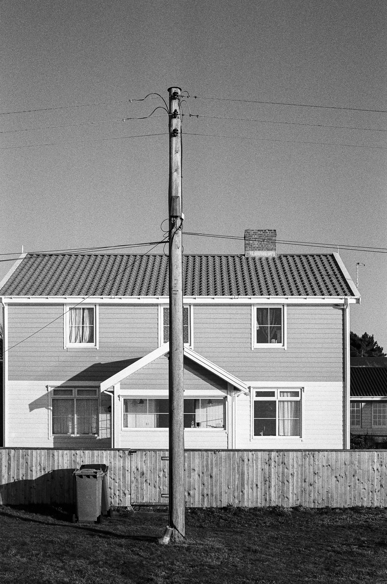 Black and white photograph of a two-story house with a sloped tiled roof, four windows on the upper floor, and a porch on the lower floor. A tall wooden utility pole with wires stands in the front yard, casting a shadow. A brick chimney is visible on