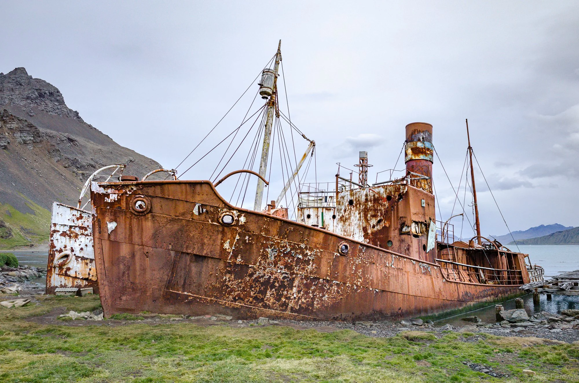 An old, rusted, abandoned ship sitting on a grassy shore with a hilly mountain in the background under cloudy sky.
