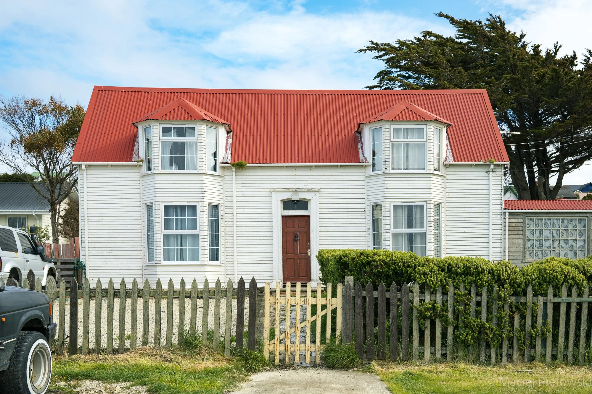 A two-story white house with a red metal roof and bay windows, surrounded by a wooden fence and green bushes. There are cars parked in front, and a large tree is in the background.