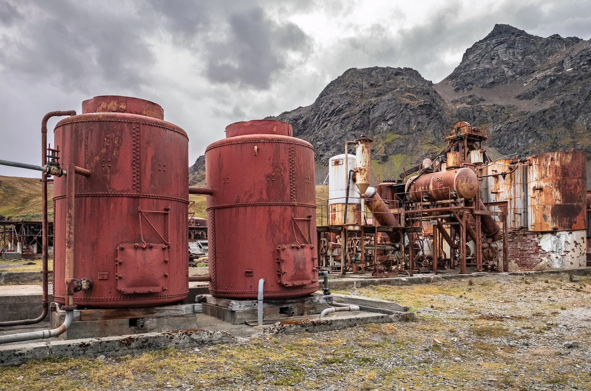 Old, rusty industrial equipment and tanks set against a mountainous landscape under cloudy skies.
