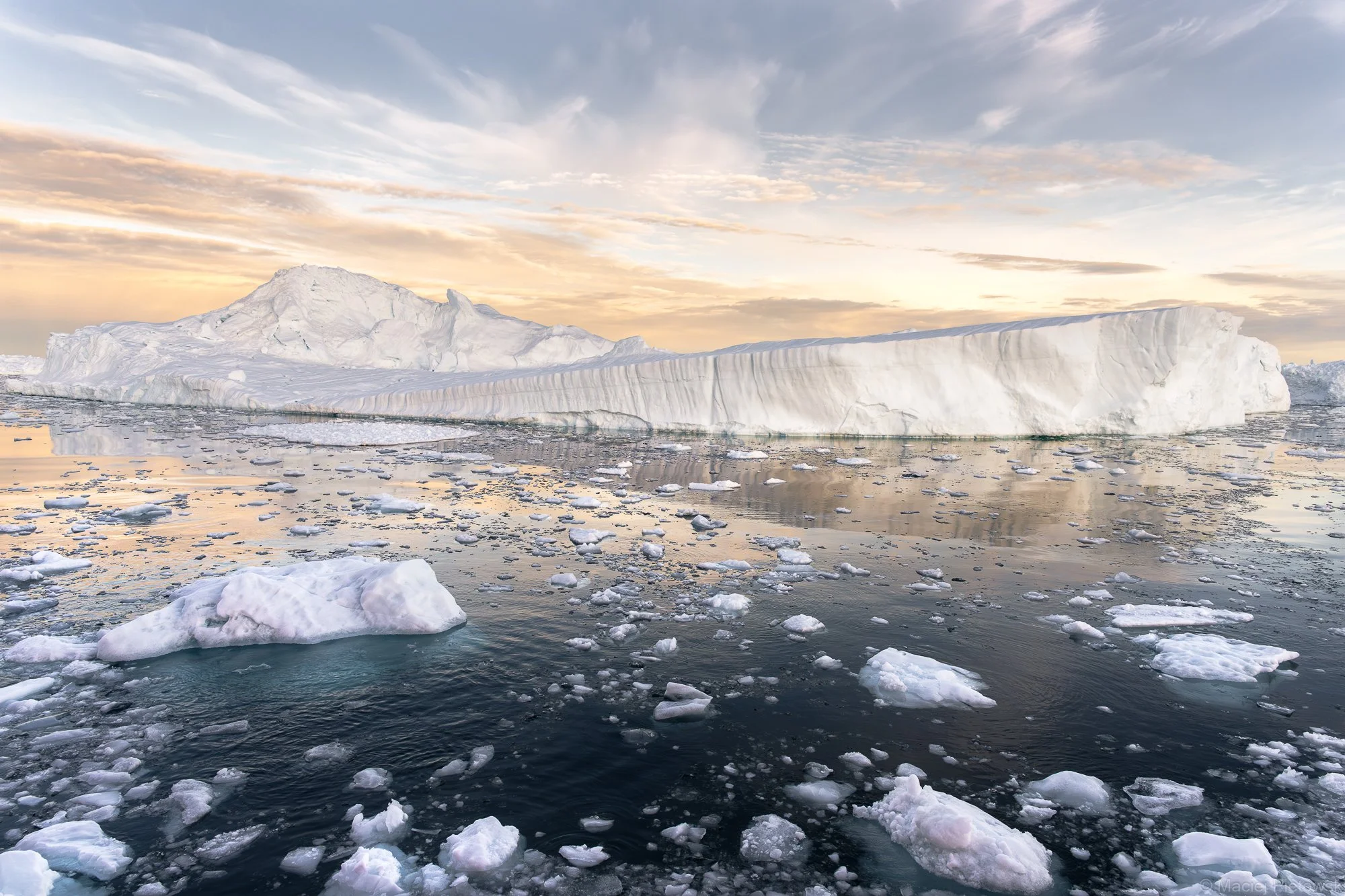 Large icy iceberg floating in Arctic waters with smaller ice chunks around, under a colorful sky during sunset or sunrise.