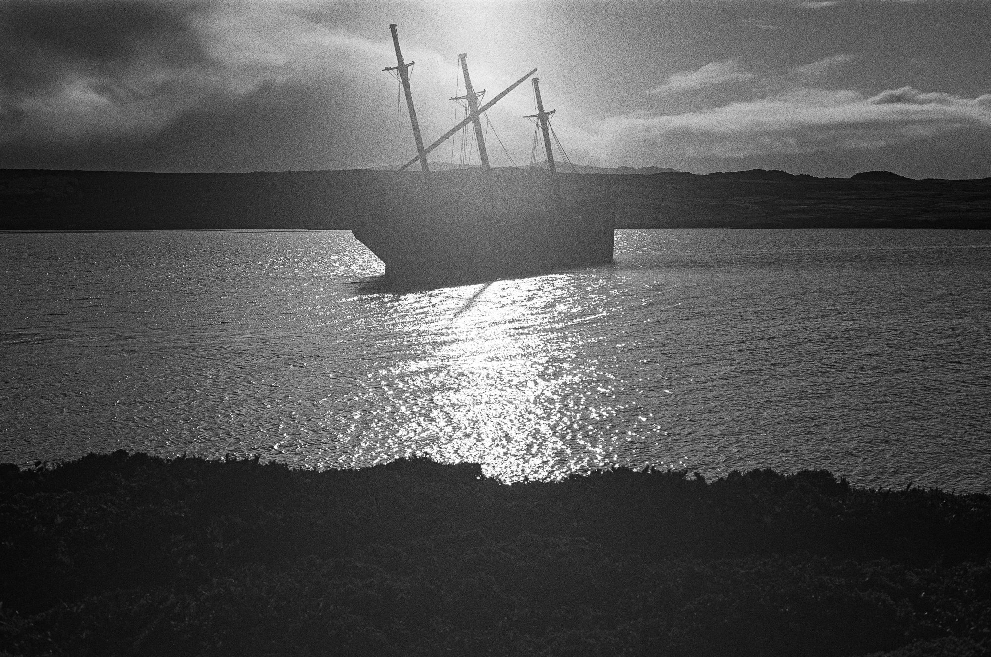 A black and white photo of a sinking ship tilted to one side in a body of water, with a hilly landscape and cloudy sky in the background.