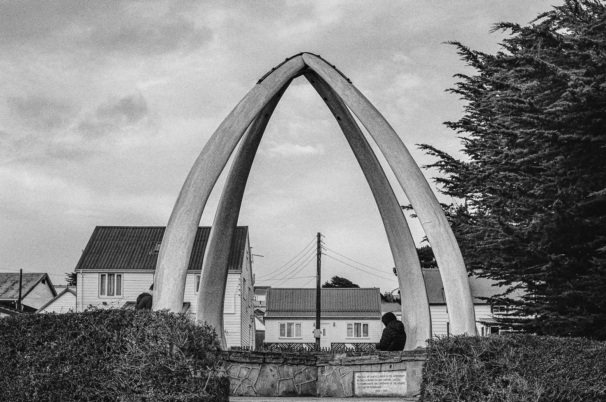 Black and white photo of a war memorial with four large, curved beams forming an arch, surrounded by houses, trees, and utility poles.