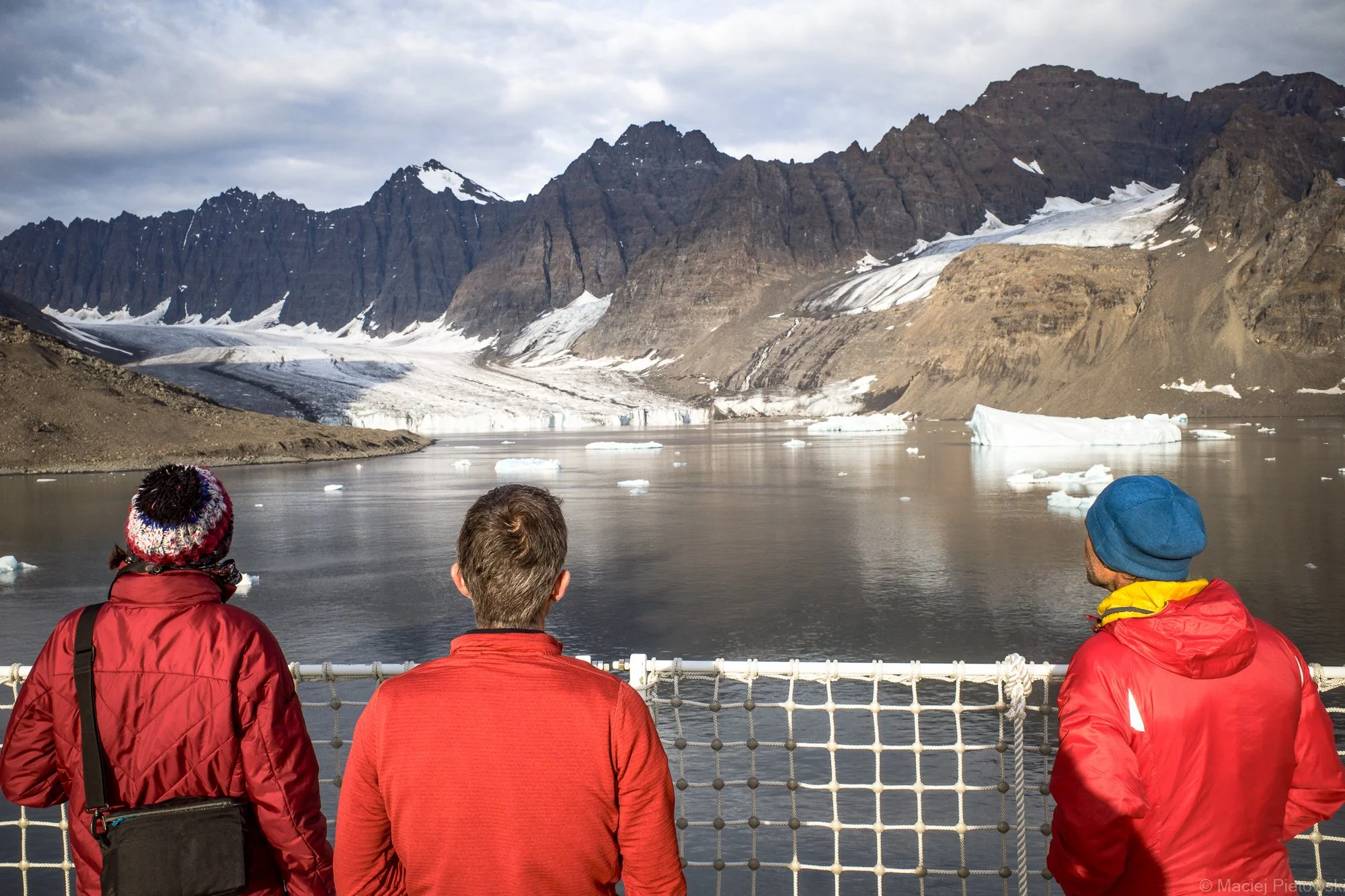 Four people wearing red and yellow jackets standing on a boat, looking at a glacier and mountain landscape with ice floating in a body of water, under cloudy sky.