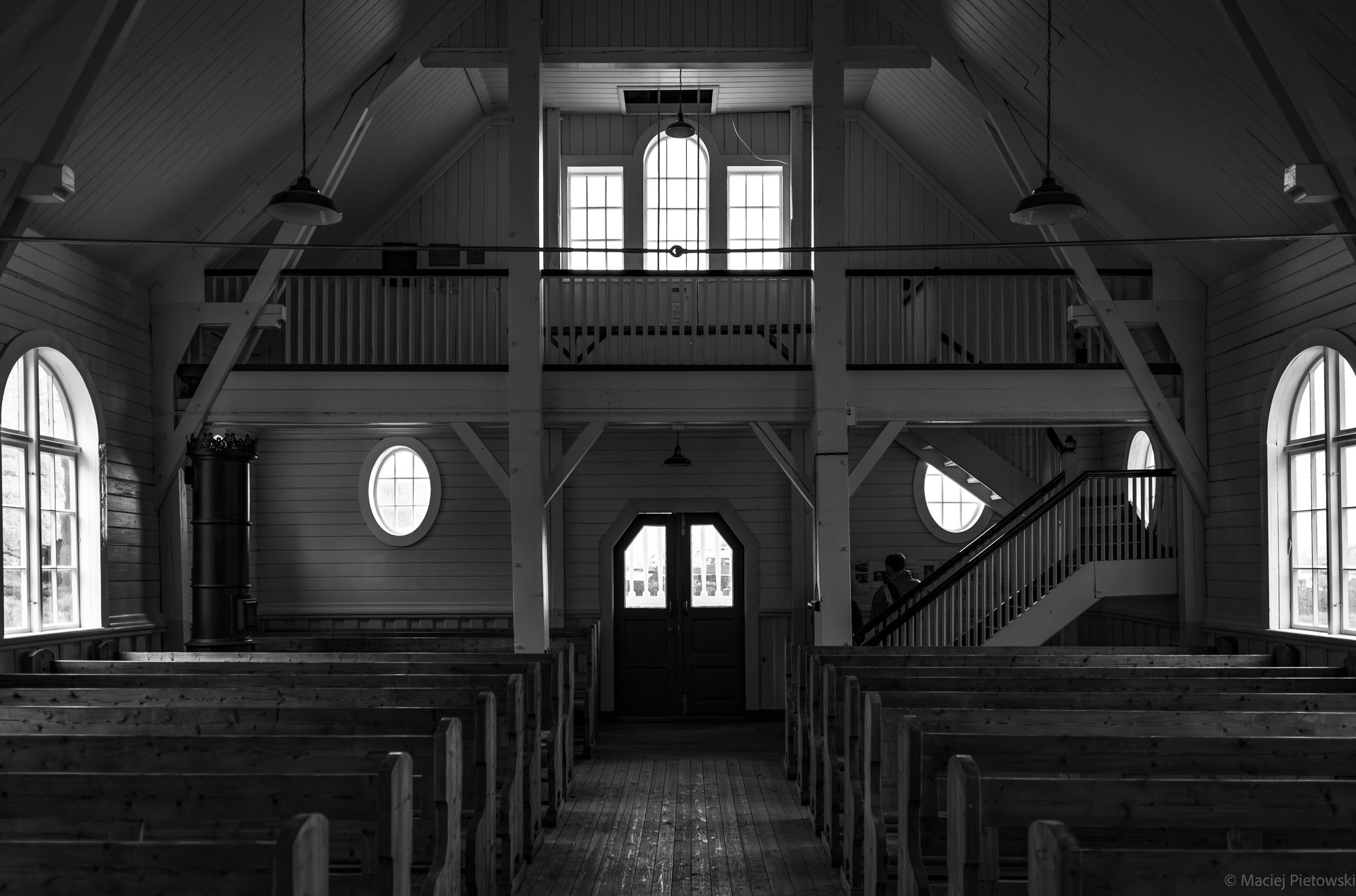 Interior of a church or auditorium with wooden beams, high windows, pews, staircase, and hanging lights, in black and white.
