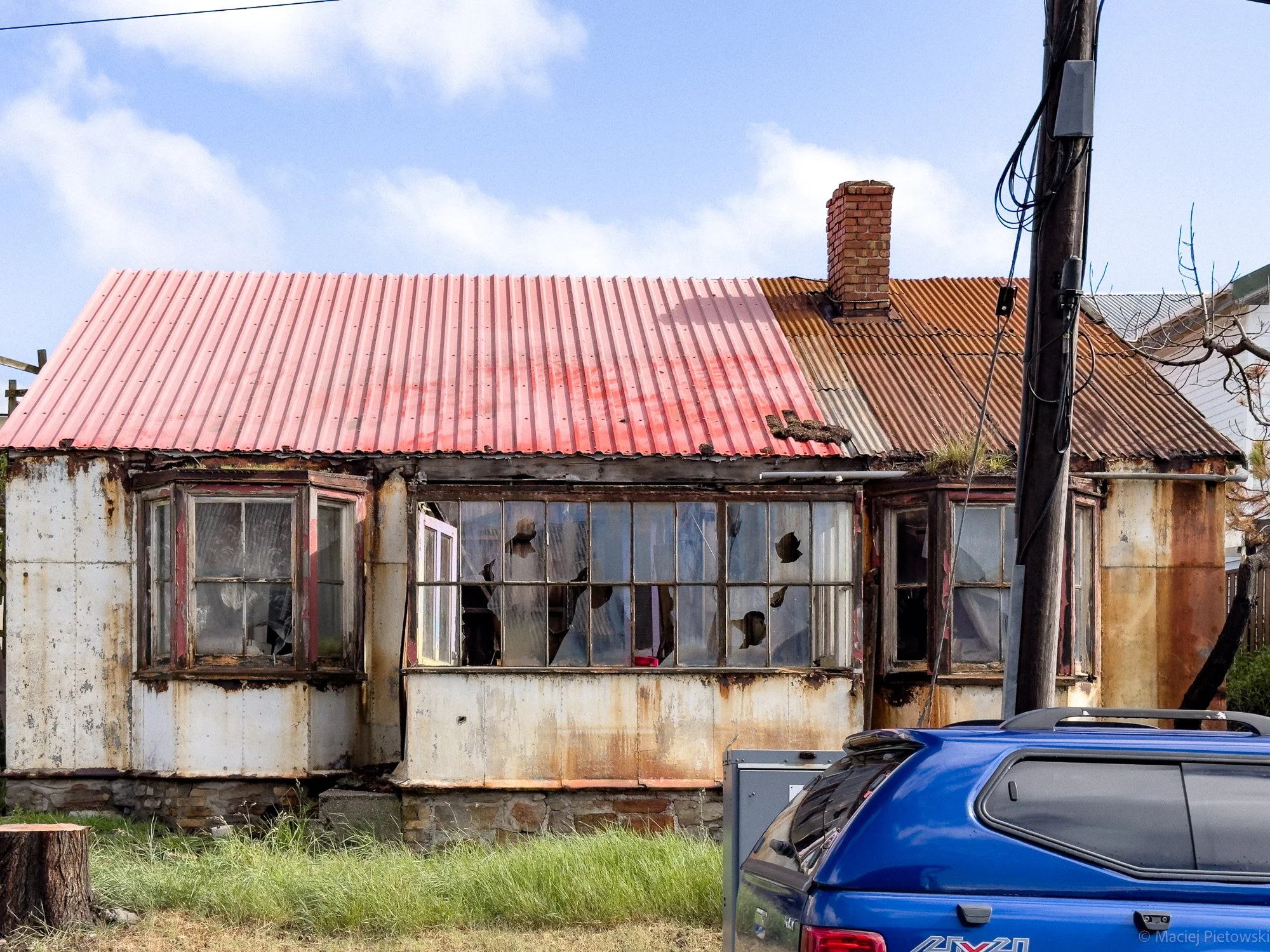 Old, weathered house with broken windows, rusty and faded red tin roof, and a brick chimney. A utility pole with wires is beside the house, and a partial view of a parked blue car is in front. The house appears abandoned and in disrepair.