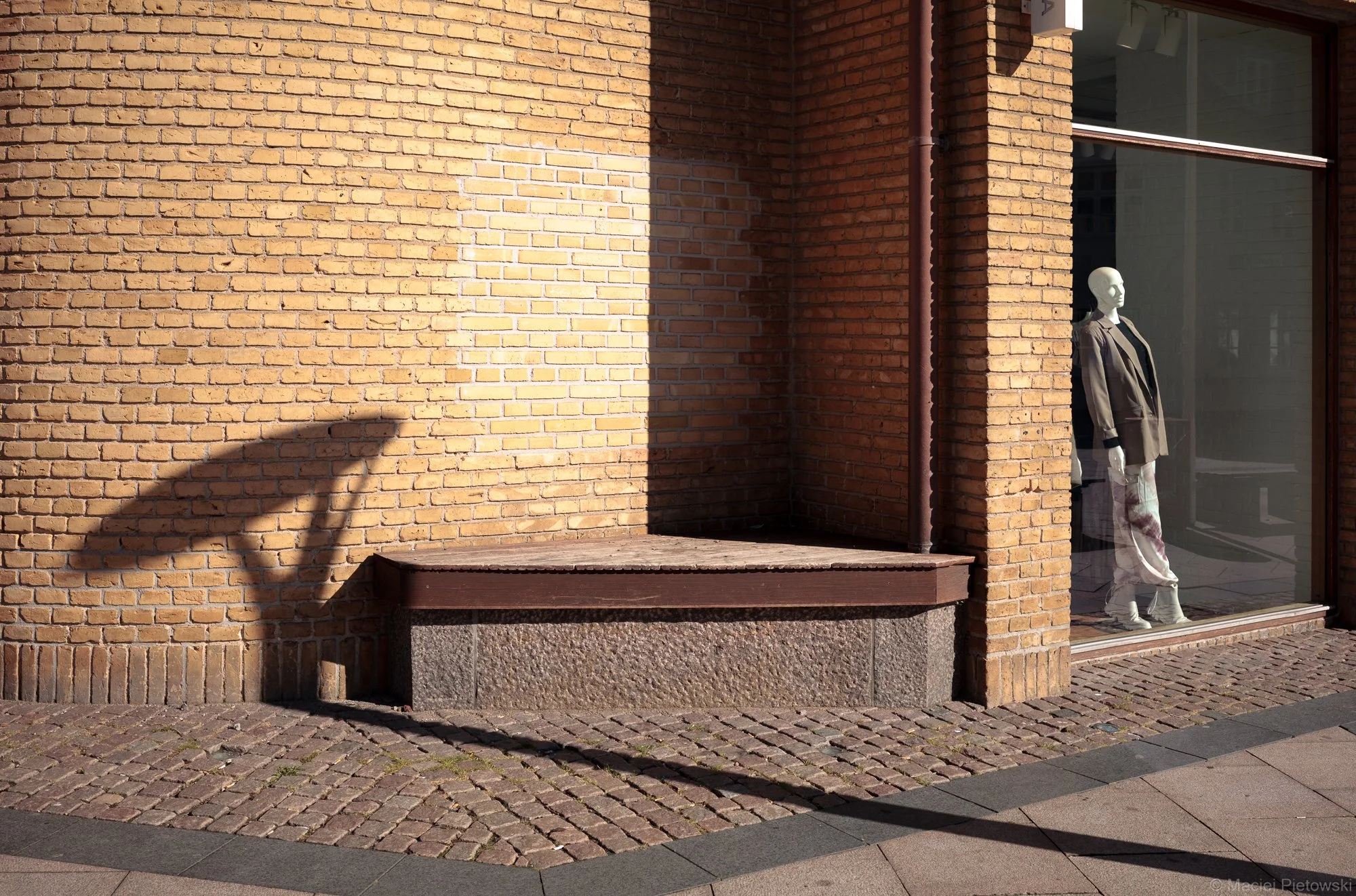 Storefront window display with a mannequin dressed in a brown jacket and beige pants, set against a brick wall exterior.
