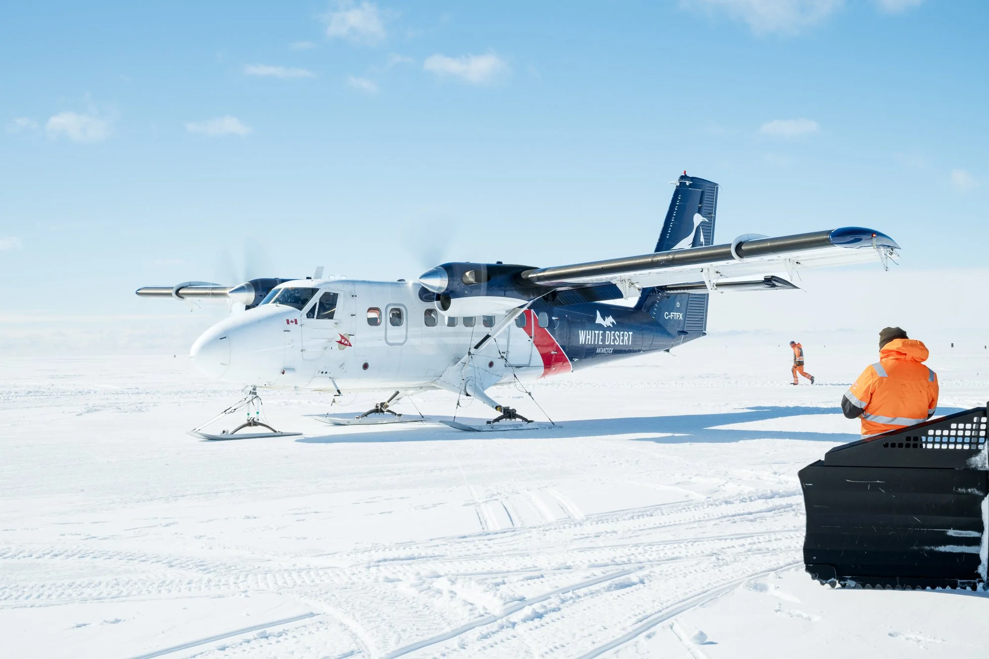A white and blue aircraft labeled 'White Desert' on a snow-covered surface, with three people in orange jackets nearby, one kneeling and two walking in the background, under a clear blue sky.