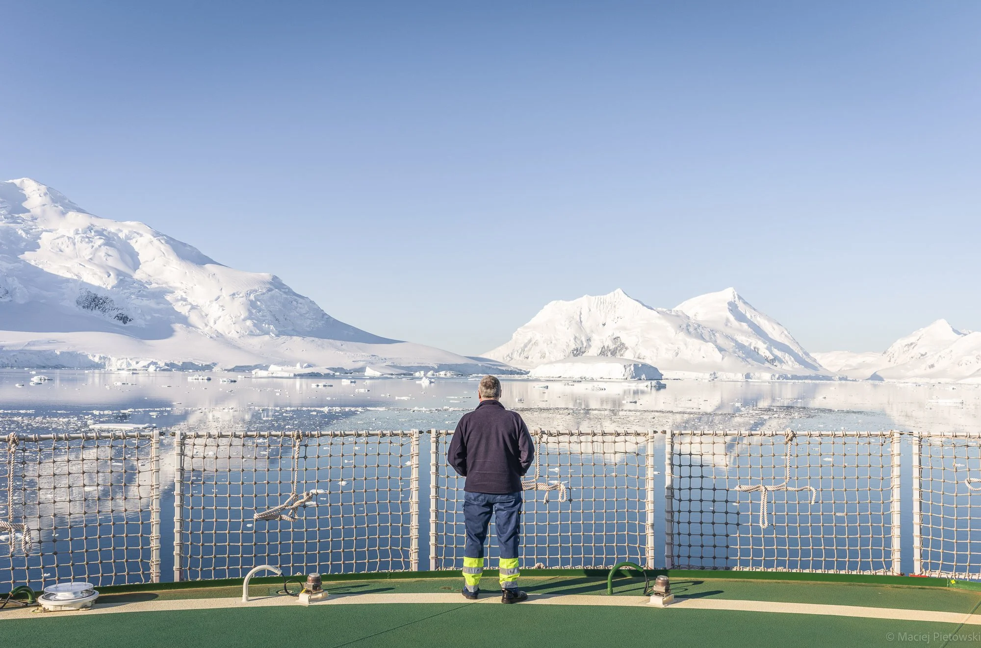 A person in a dark jacket and yellow reflective pants stands on a ship deck looking at icy mountains and water in a cold, snowy landscape. A safety net is in the foreground.