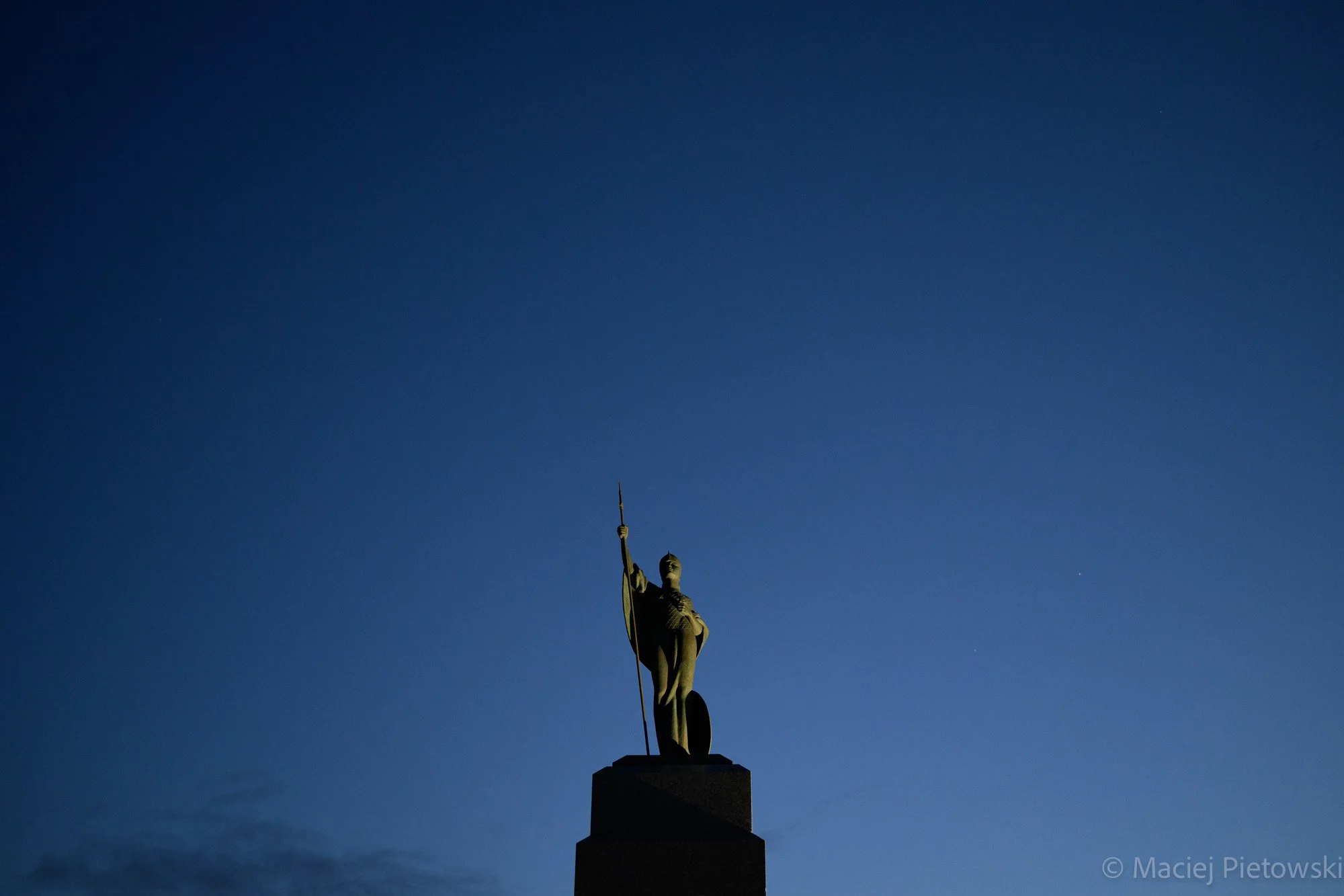 Statue of a historical figure holding a staff, standing on a pedestal against a dark blue sky.