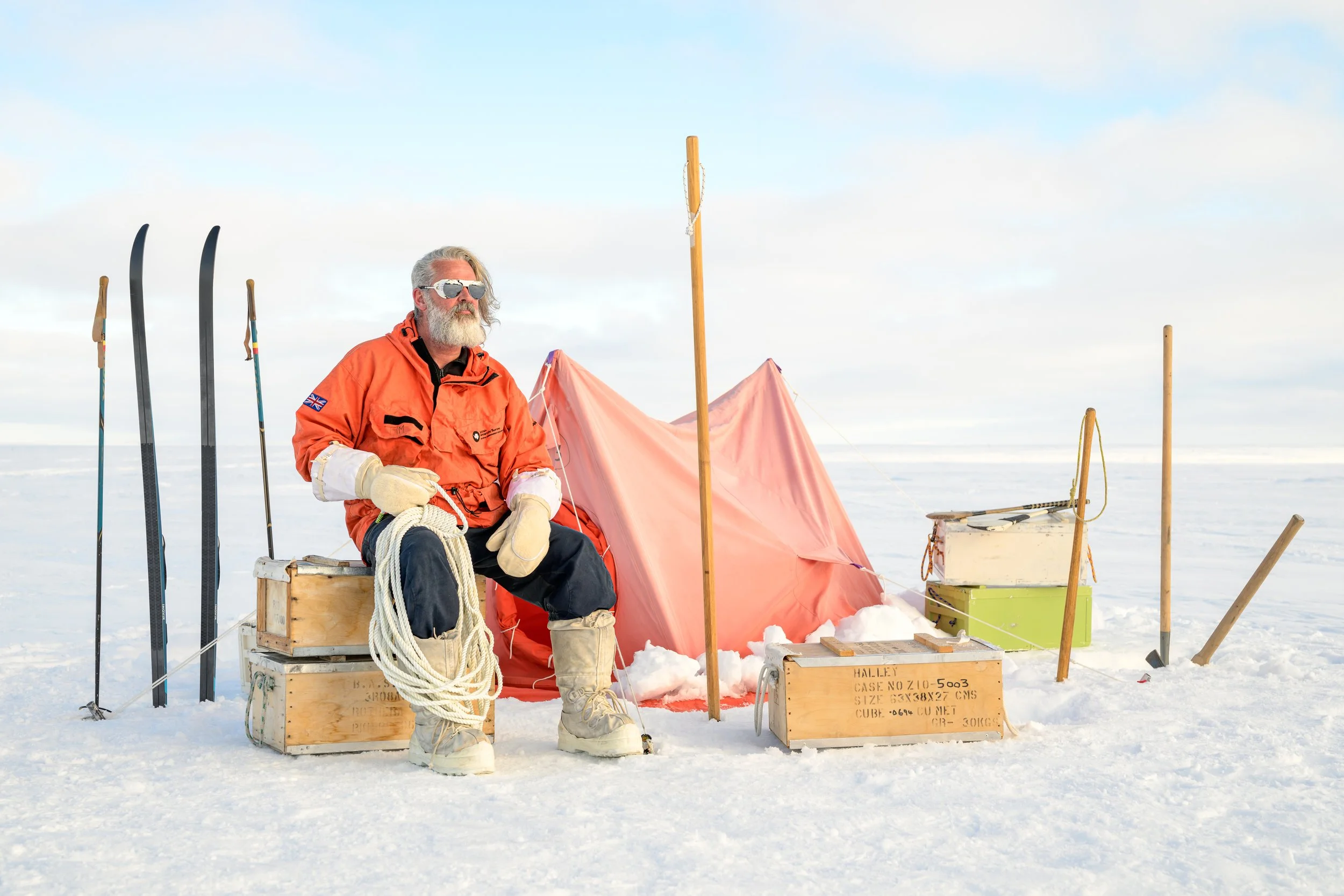 A bearded man wearing sunglasses and an orange survival suit sitting on a wooden box in a snowy landscape, surrounded by equipment and flags, with skis and a tent in the background.