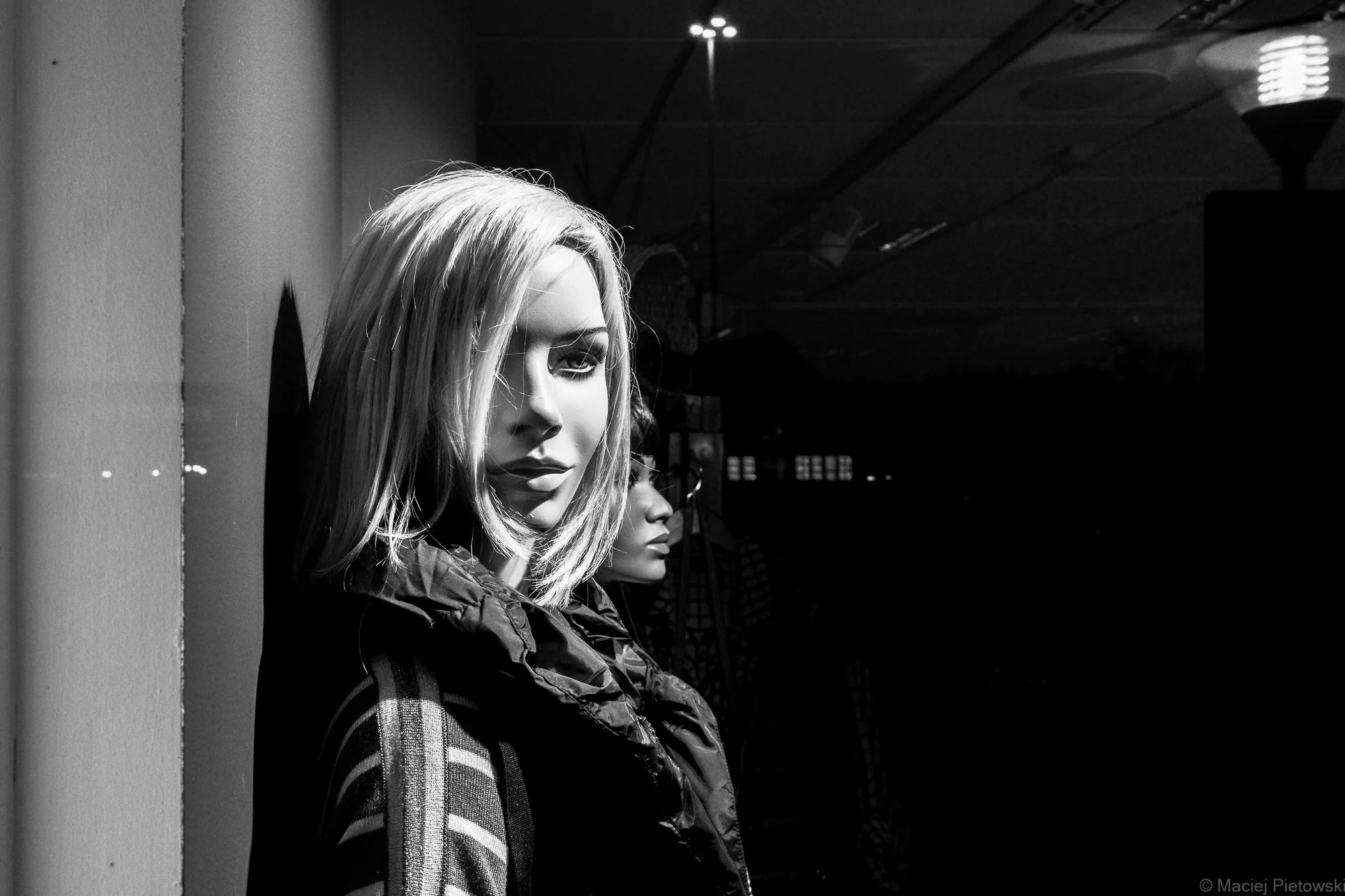 Black and white photo of a mannequin with shoulder-length blonde hair, wearing a jacket, positioned against a wall with a slightly darker background.