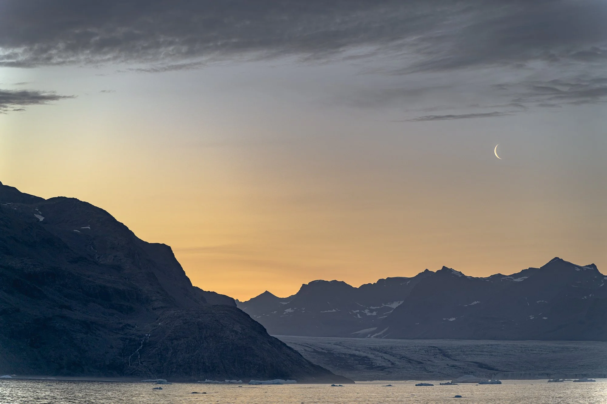 A mountainous landscape at dusk, with a crescent moon in the sky. The mountains are rugged with some patches of snow and the sky is partly cloudy with a gradient of colors from yellow to dark gray.