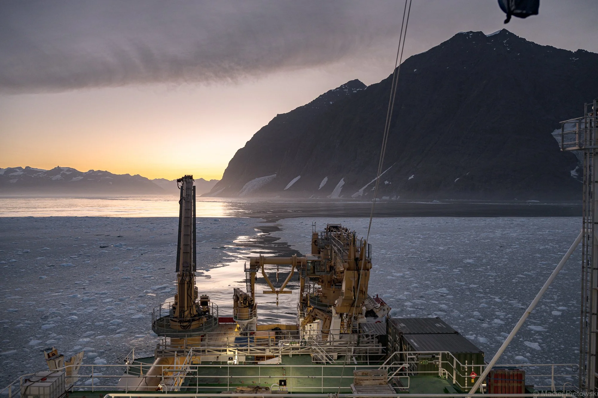 View from a ship's deck showing a snowy landscape with mountains in the distance, overcast sky, and icy waters with floating ice chunks, in a cold, remote environment.