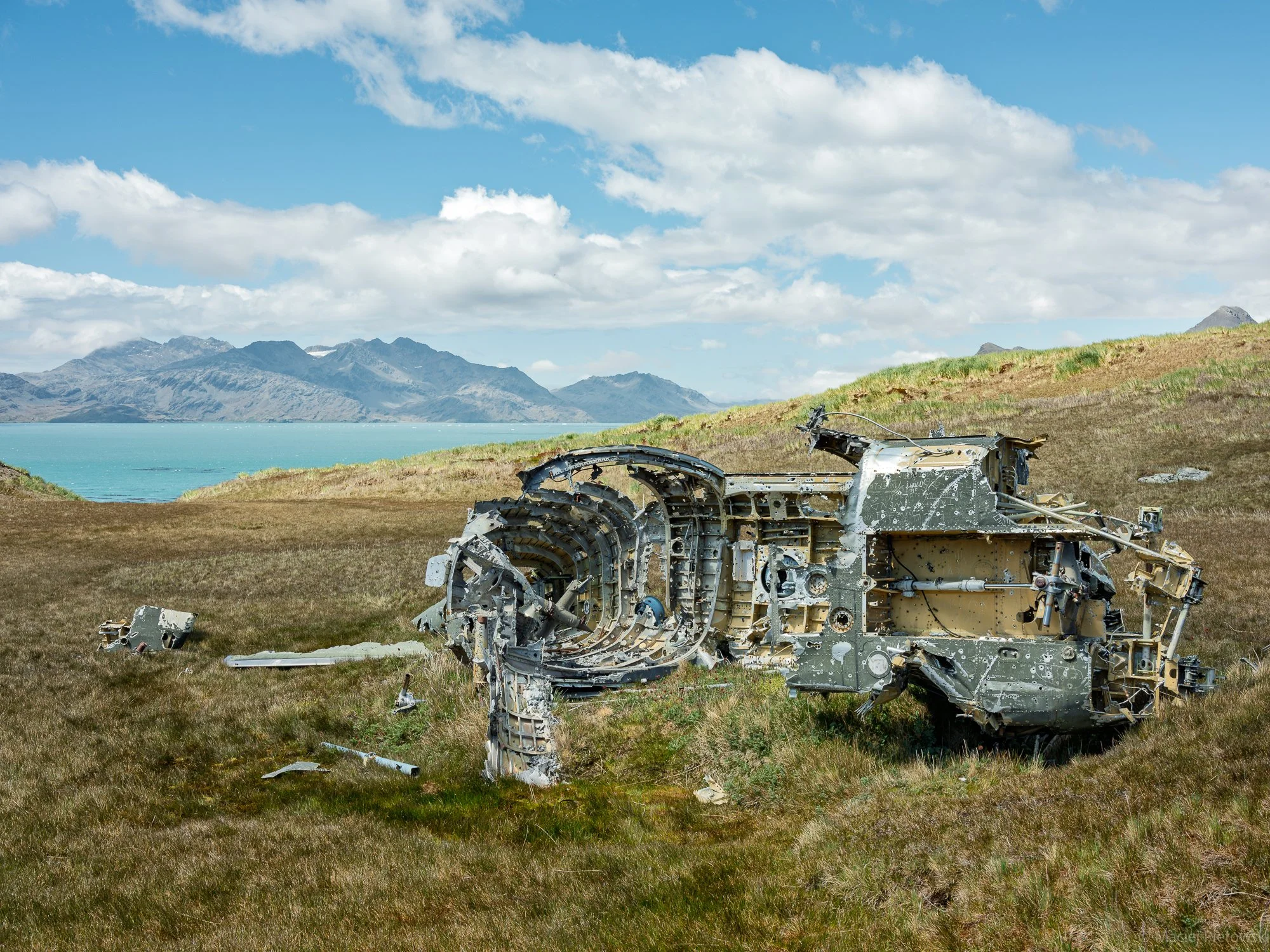 A landscape with mountains and a lake in the background, and the wreckage of a destroyed vehicle or spacecraft in the foreground on grassy terrain.