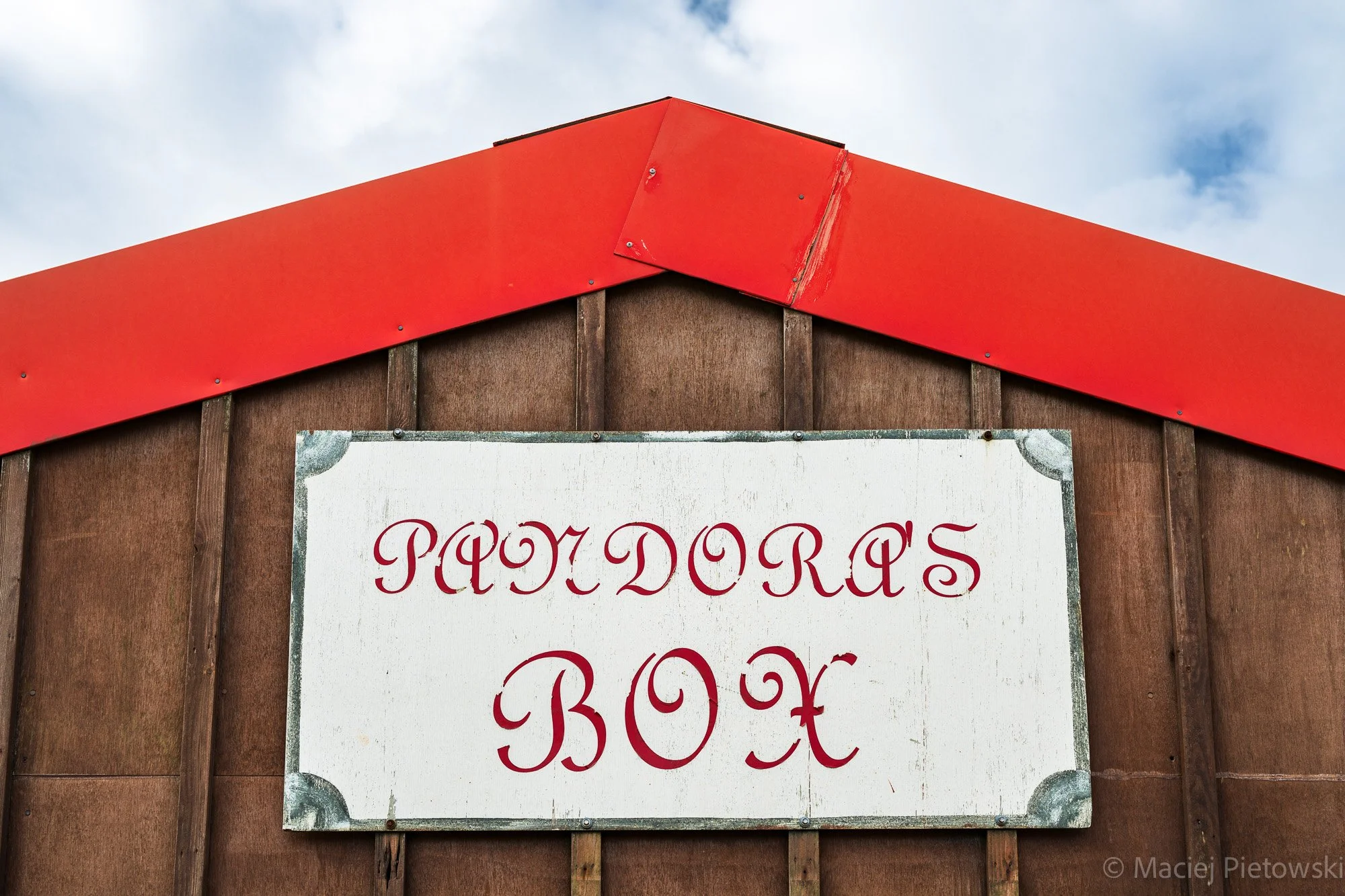 Close-up of a sign with red cursive text on a white background, attached to a wooden structure with a red gable roof, against a cloudy sky.