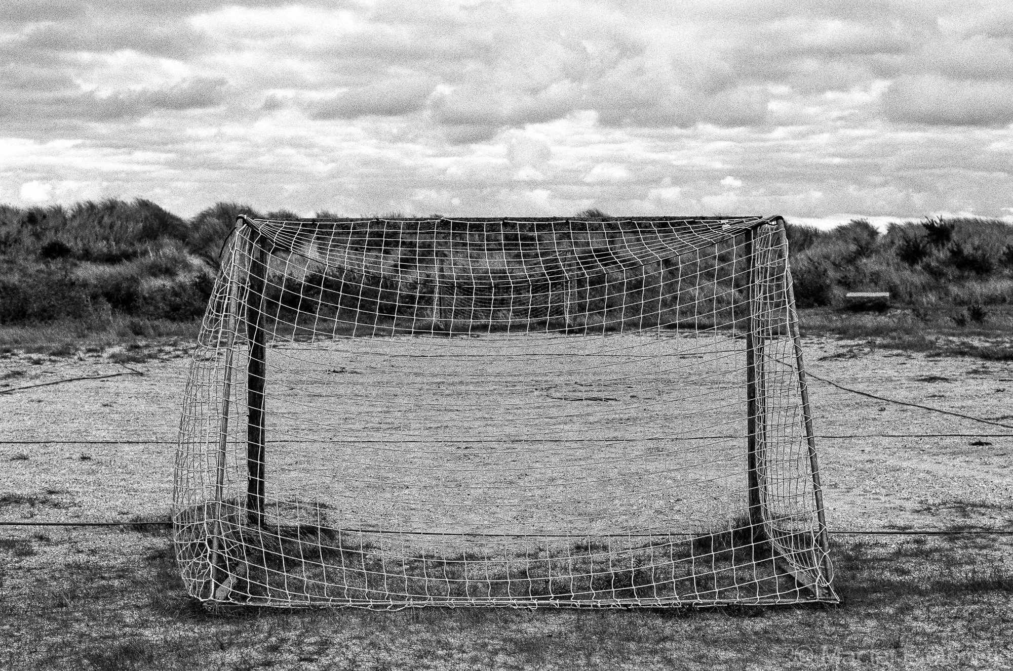 A soccer goal on a sandy, open field with bushes and cloudy sky in the background, all depicted in black and white.