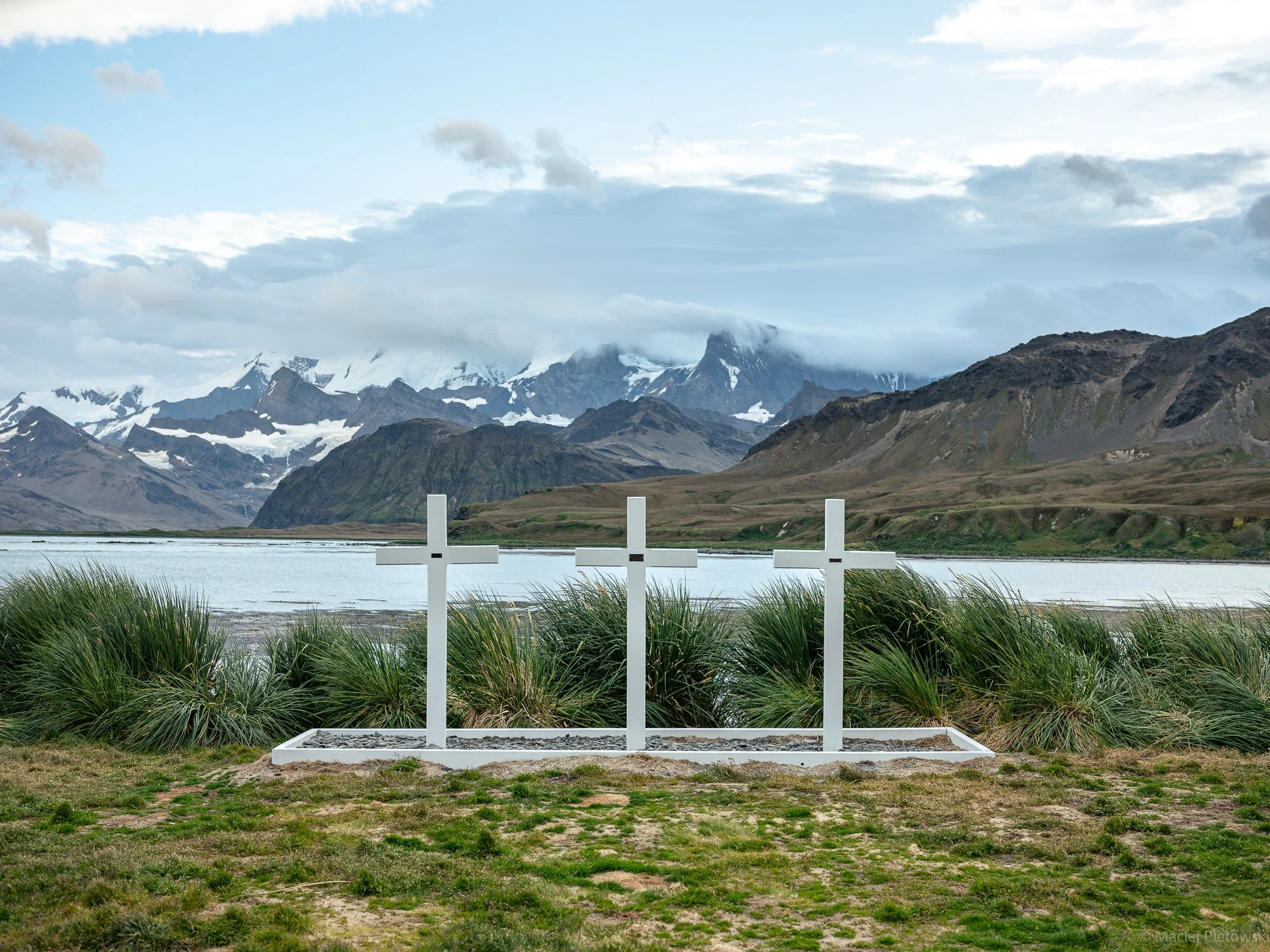 Memorial crosses placed by a lake with mountains in the background, overcast sky.
