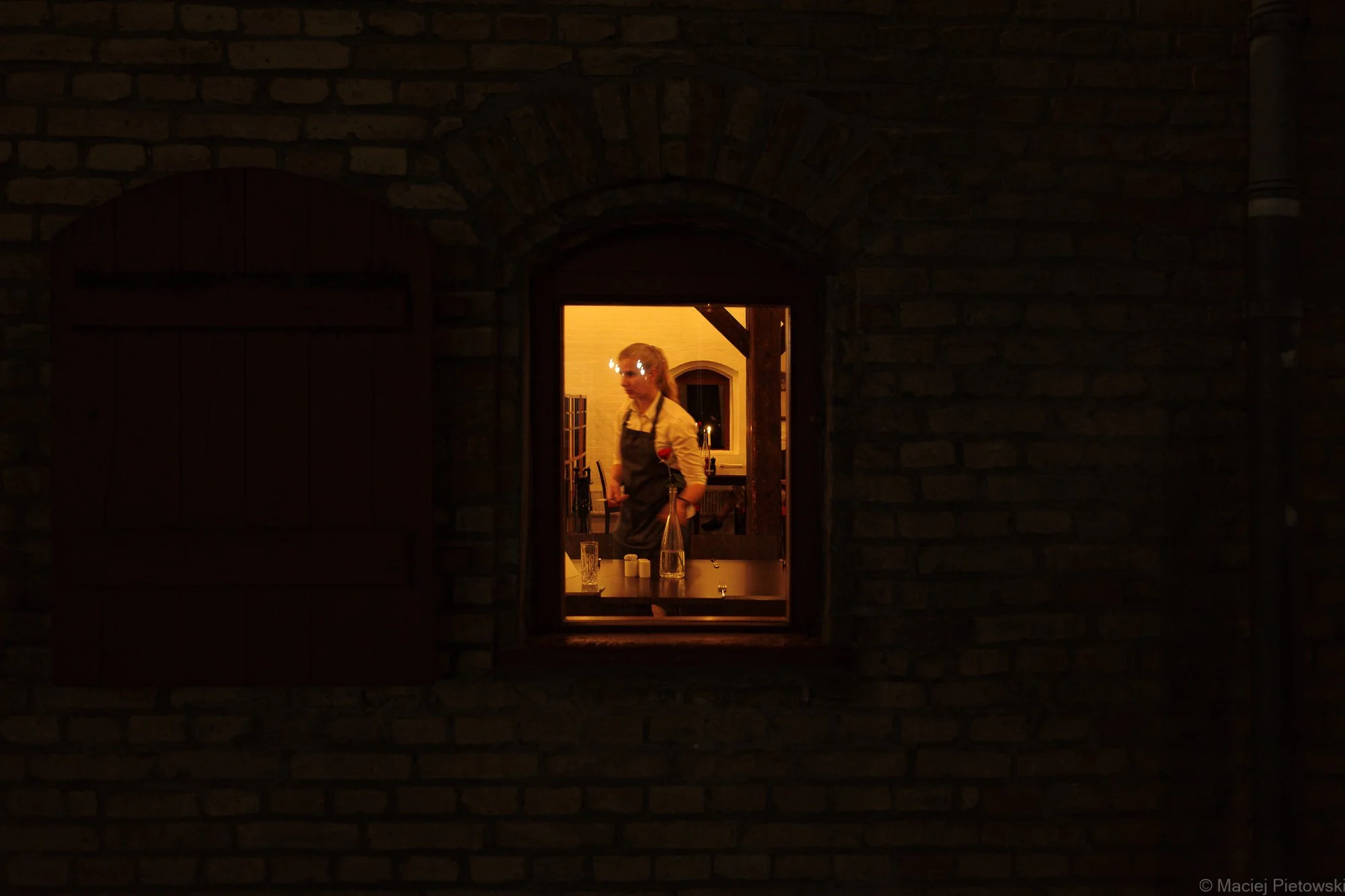 A person inside a well-lit kitchen viewed through a small window in a dark brick wall.